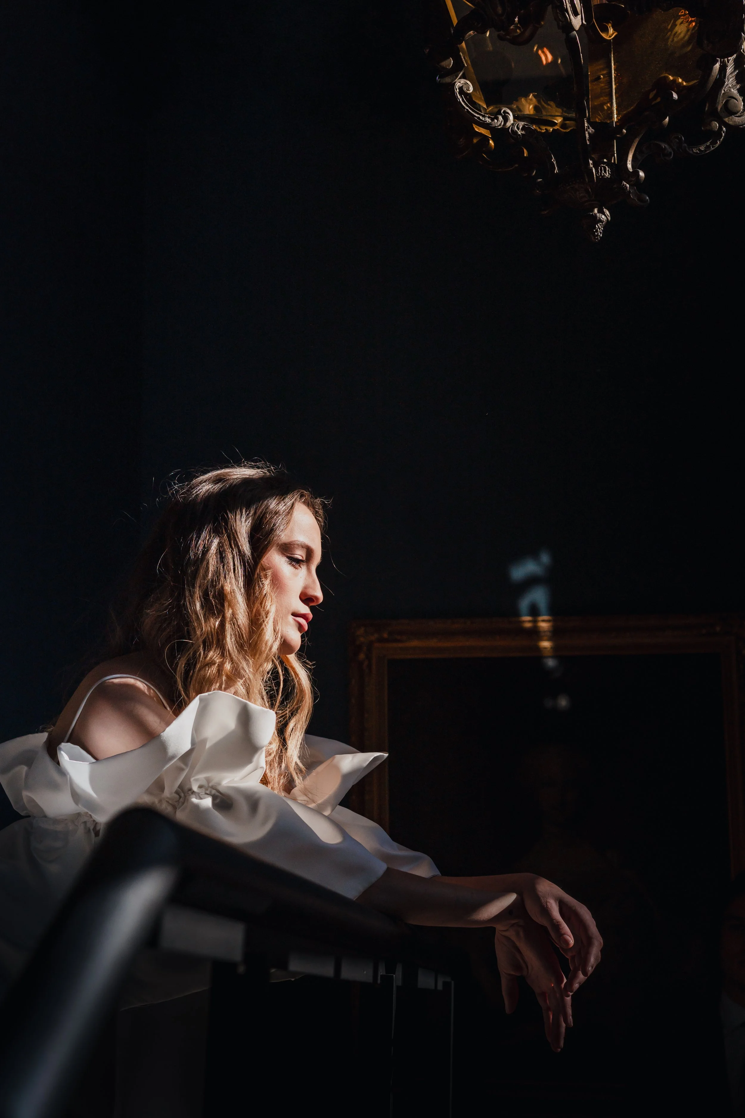 A woman with wavy blonde hair in a white, ruffled dress, standing inside a dimly lit room with dark walls, leaning on a railing, with a chandelier above.
