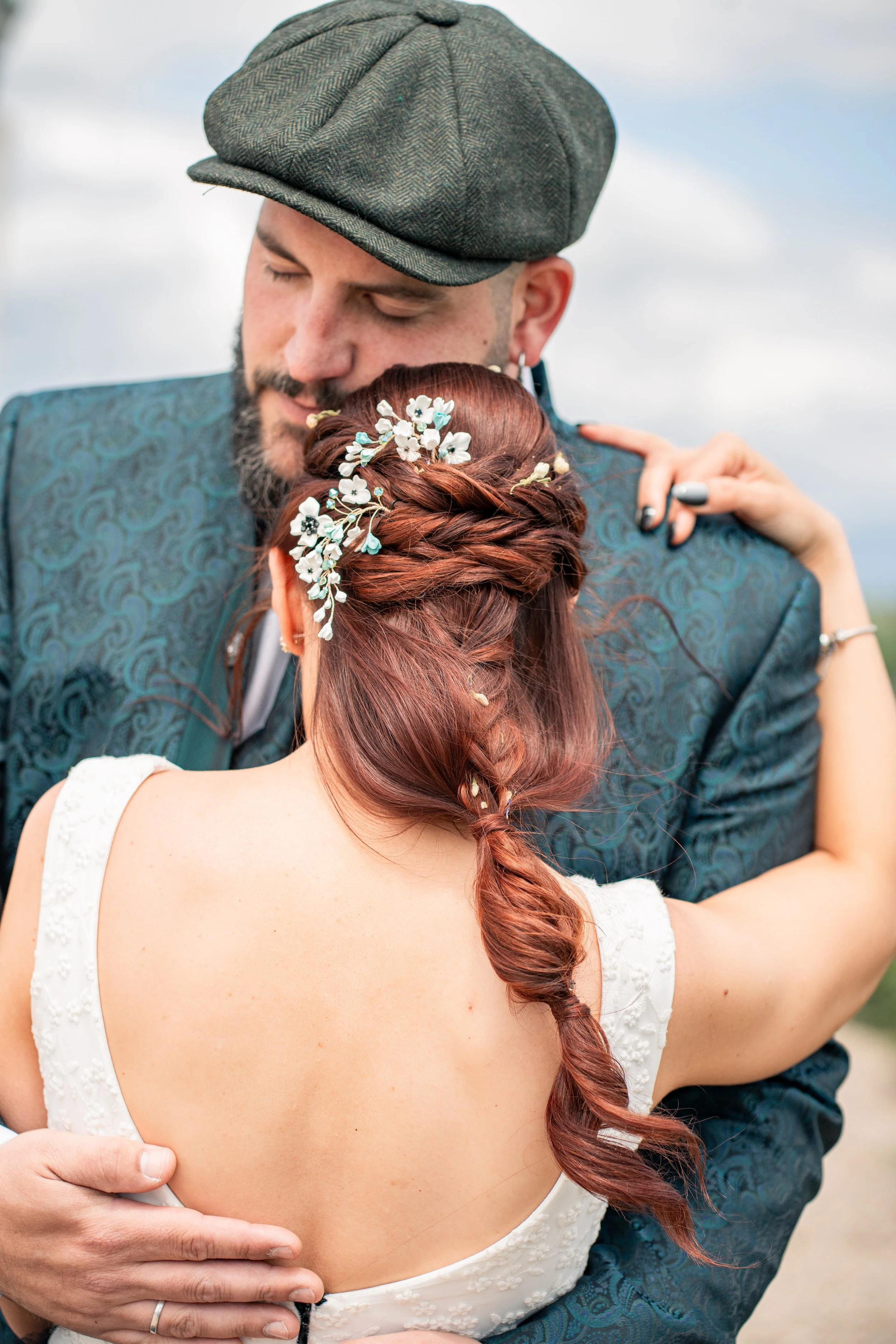 A man and woman embrace, with the woman's back to the camera. The woman has long, styled red hair with a floral hairpiece, and the man wears a gray hat and a dark, patterned suit jacket. They are outdoors against a cloudy sky.
