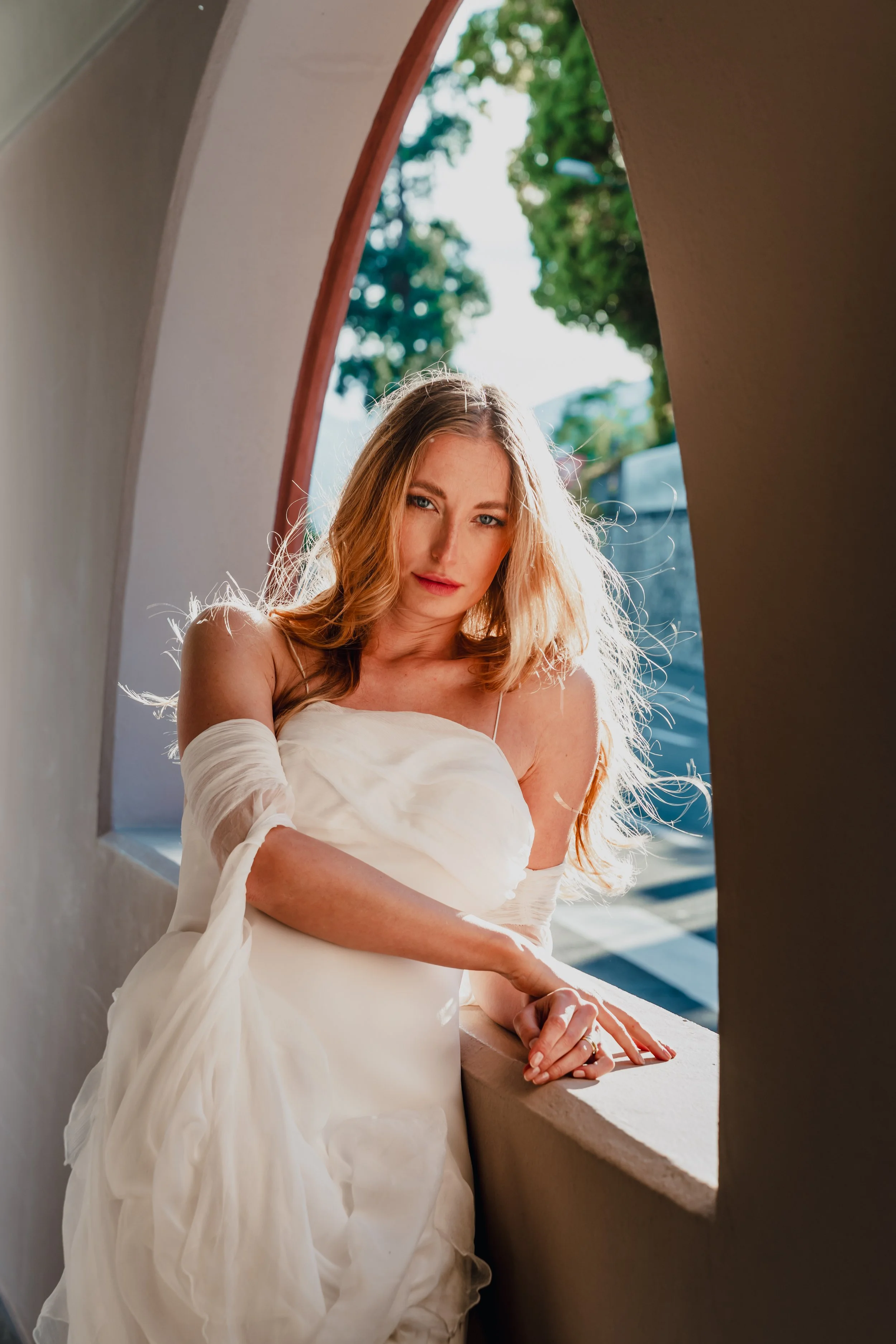 A woman with long, wavy hair in a white dress leaning on a curved windowsill of a beige-colored building, with sunlight illuminating her face and hair and trees visible outside.