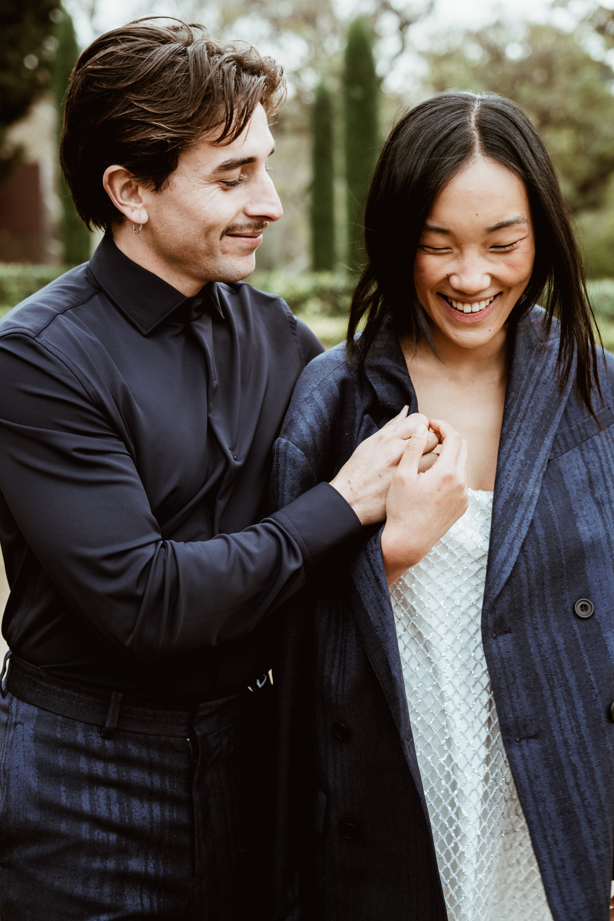 A man with brown hair, wearing a black shirt, is smiling and touching a woman with black hair, wearing a blue blazer, who is also smiling and looking down.