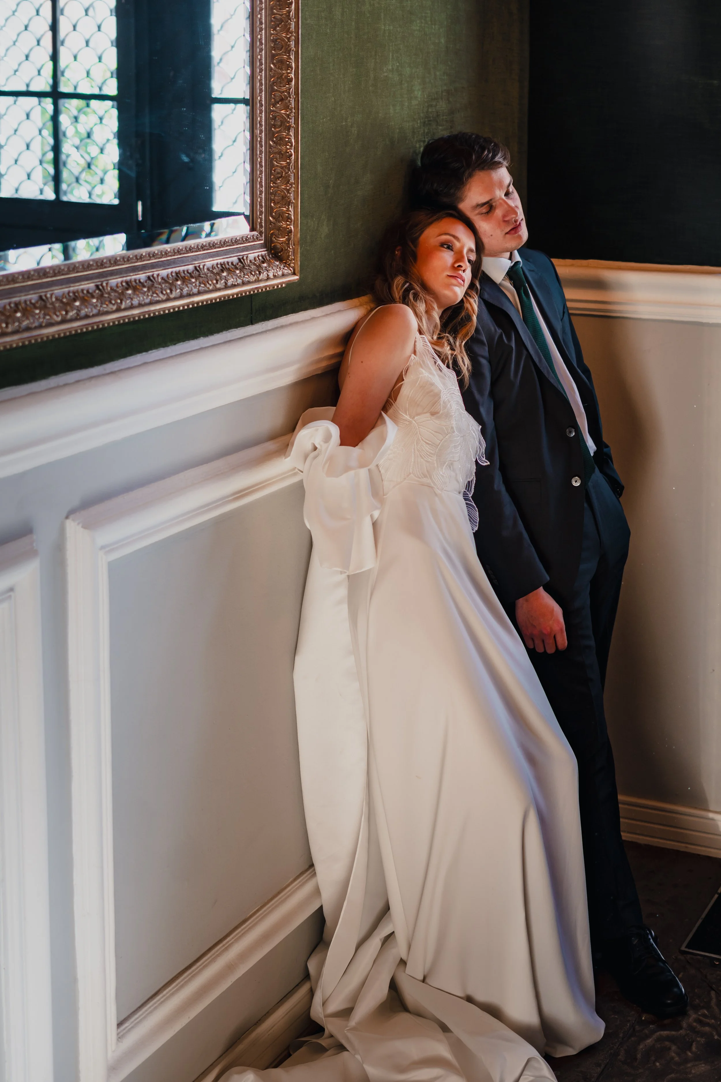 A bride and groom lean against a wall in a dimly lit room, appearing tired or contemplative, with the bride in a white gown and the groom in a dark suit.