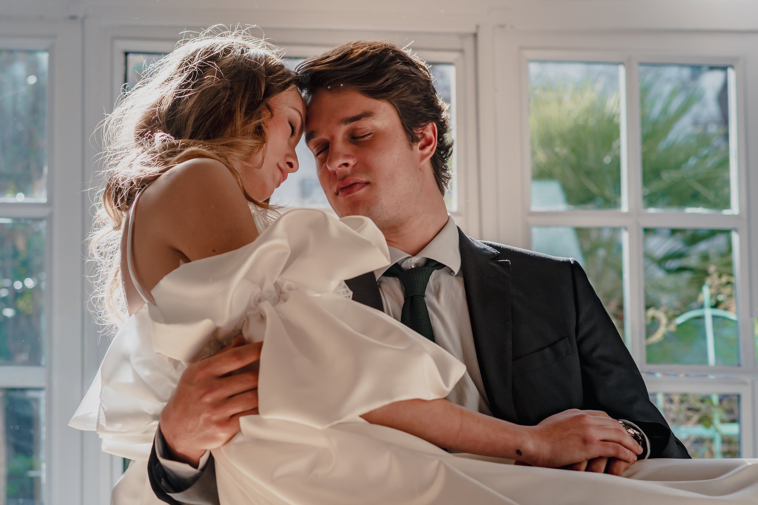 A man in a suit holding a woman in a white dress with a large bow, both with eyes closed, leaning their foreheads together, in front of a large window with plants outside.