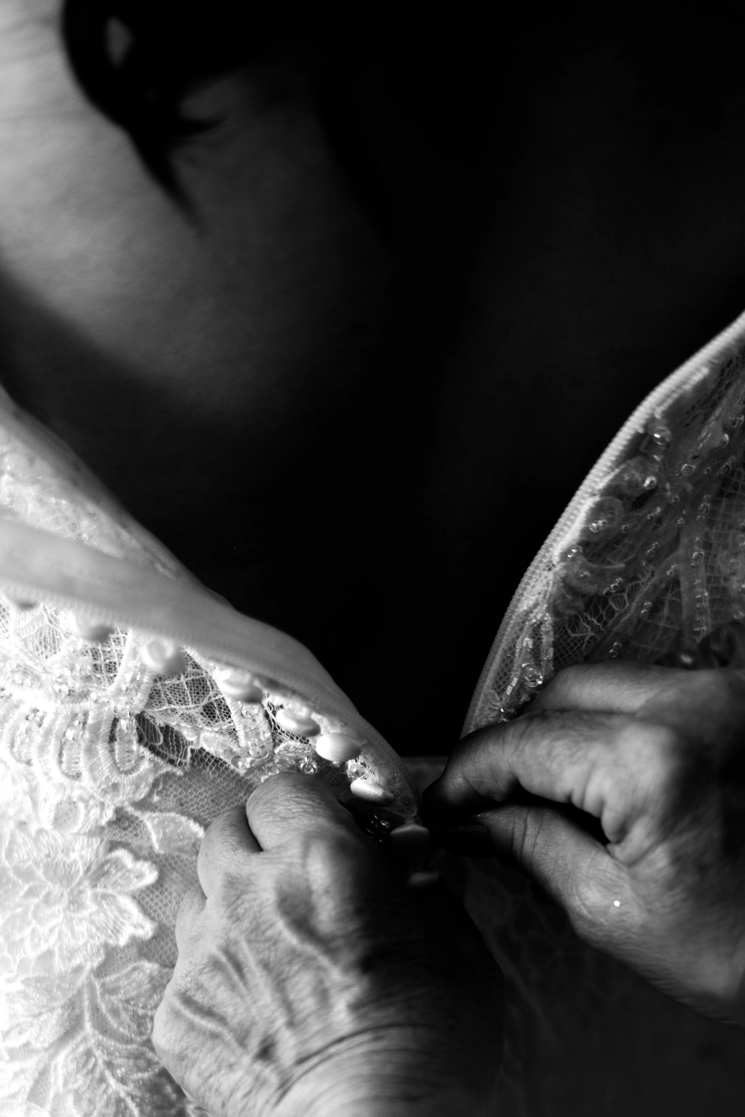 Close-up of a person buttoning a lace and sequined garment, showing their hands and part of their face in black and white.