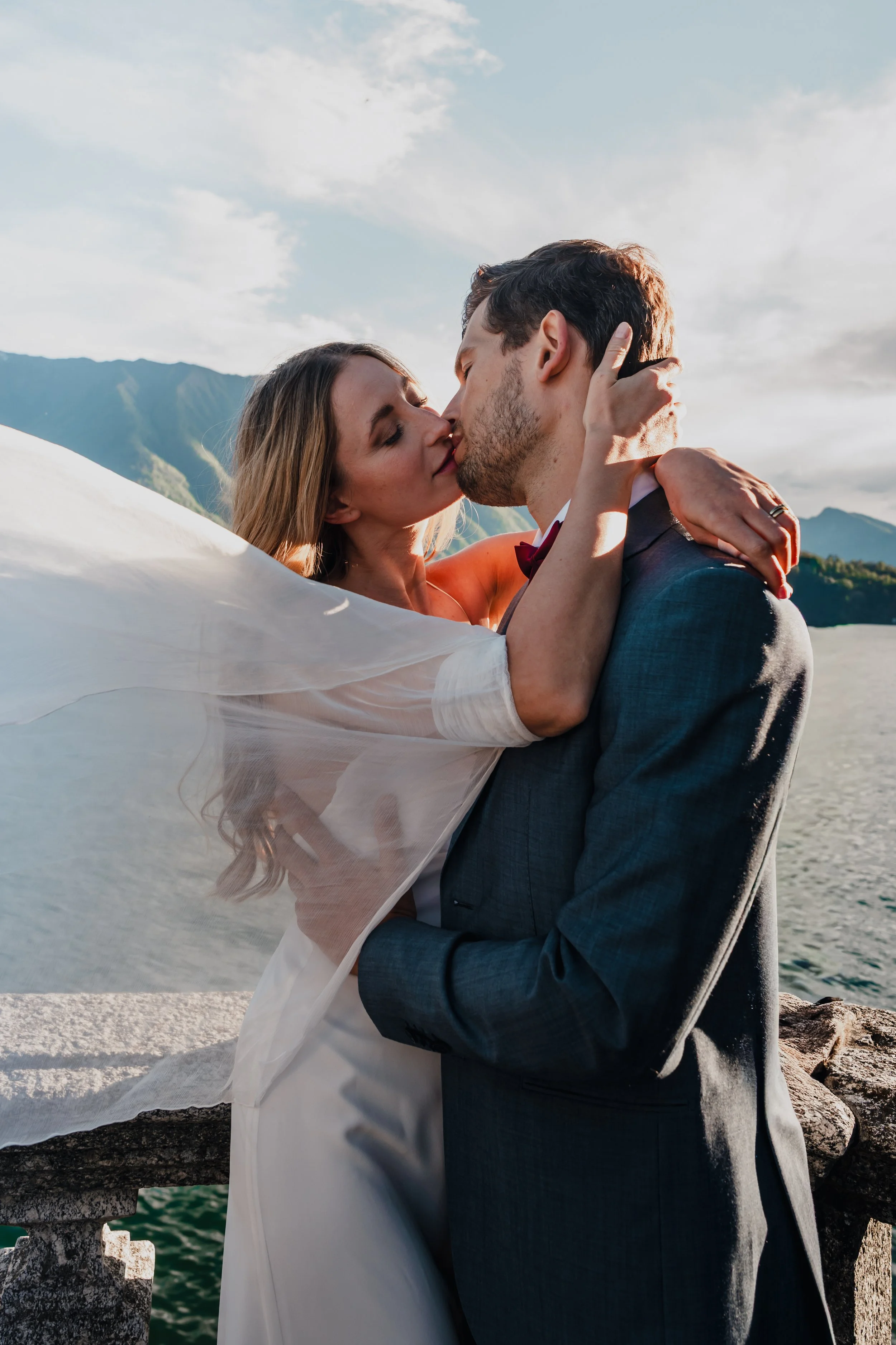 A newlywed couple sharing a kiss outdoors with a mountain and lake background, the woman in a wedding dress and a veil, the man in a dark suit.