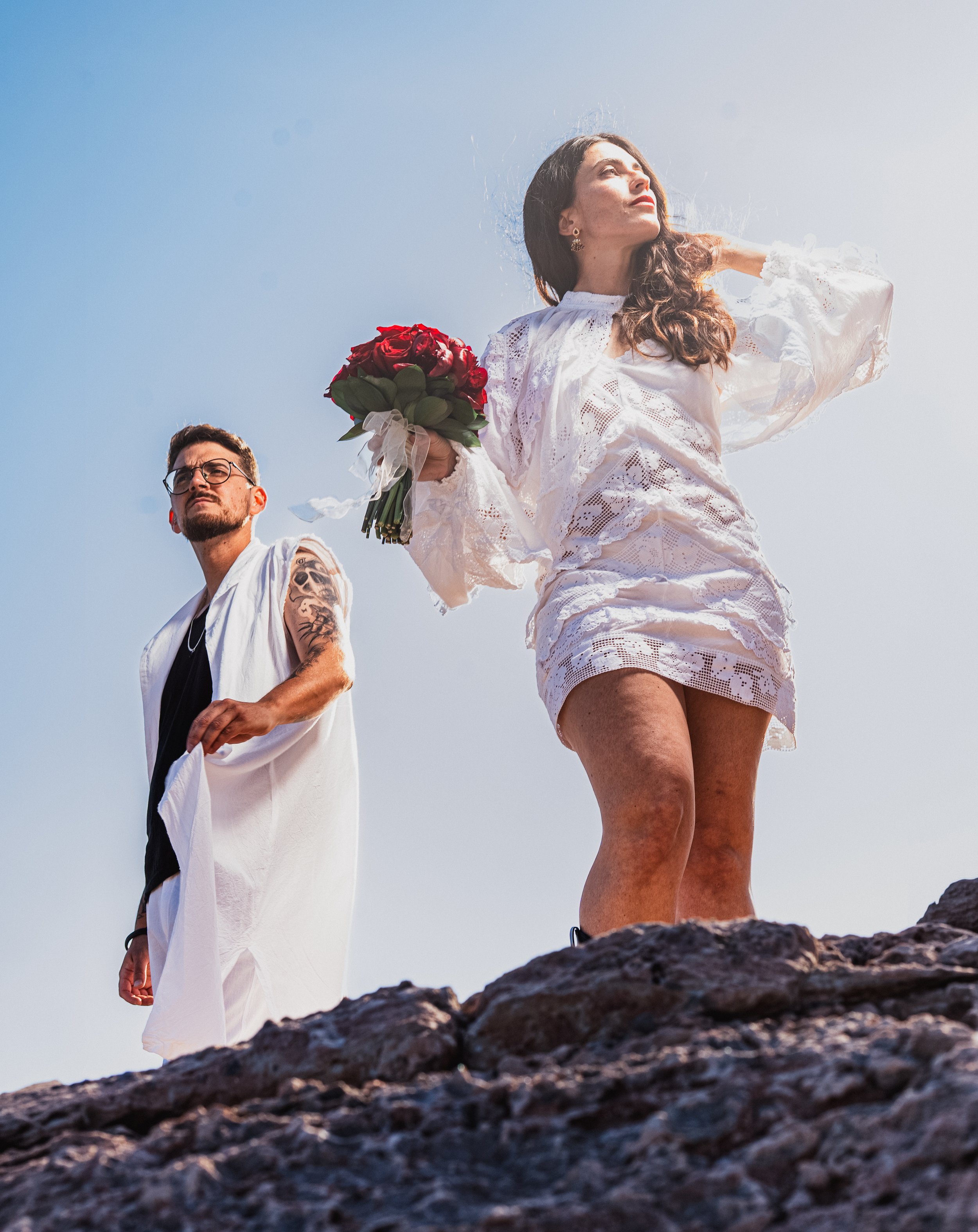 Mujer sosteniendo ramo de rosas rojas en un entorno al aire libre, otra persona acompañándola, ambos de pie sobre rocas, con cielo despejado de fondo.