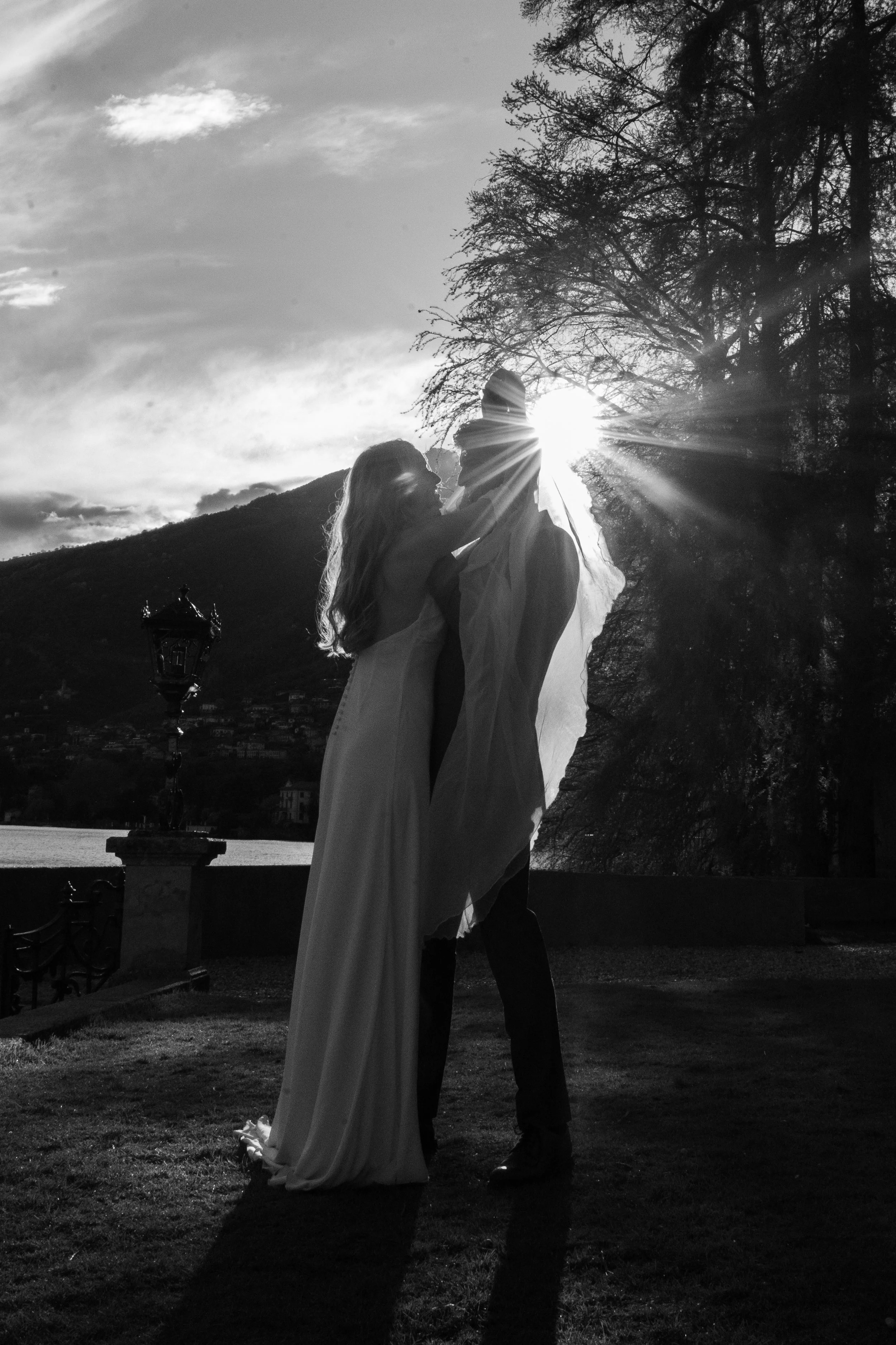A black and white photo of a couple kissing outdoors during sunset, with the sun behind them creating a silhouette effect, trees, and a body of water in the background.