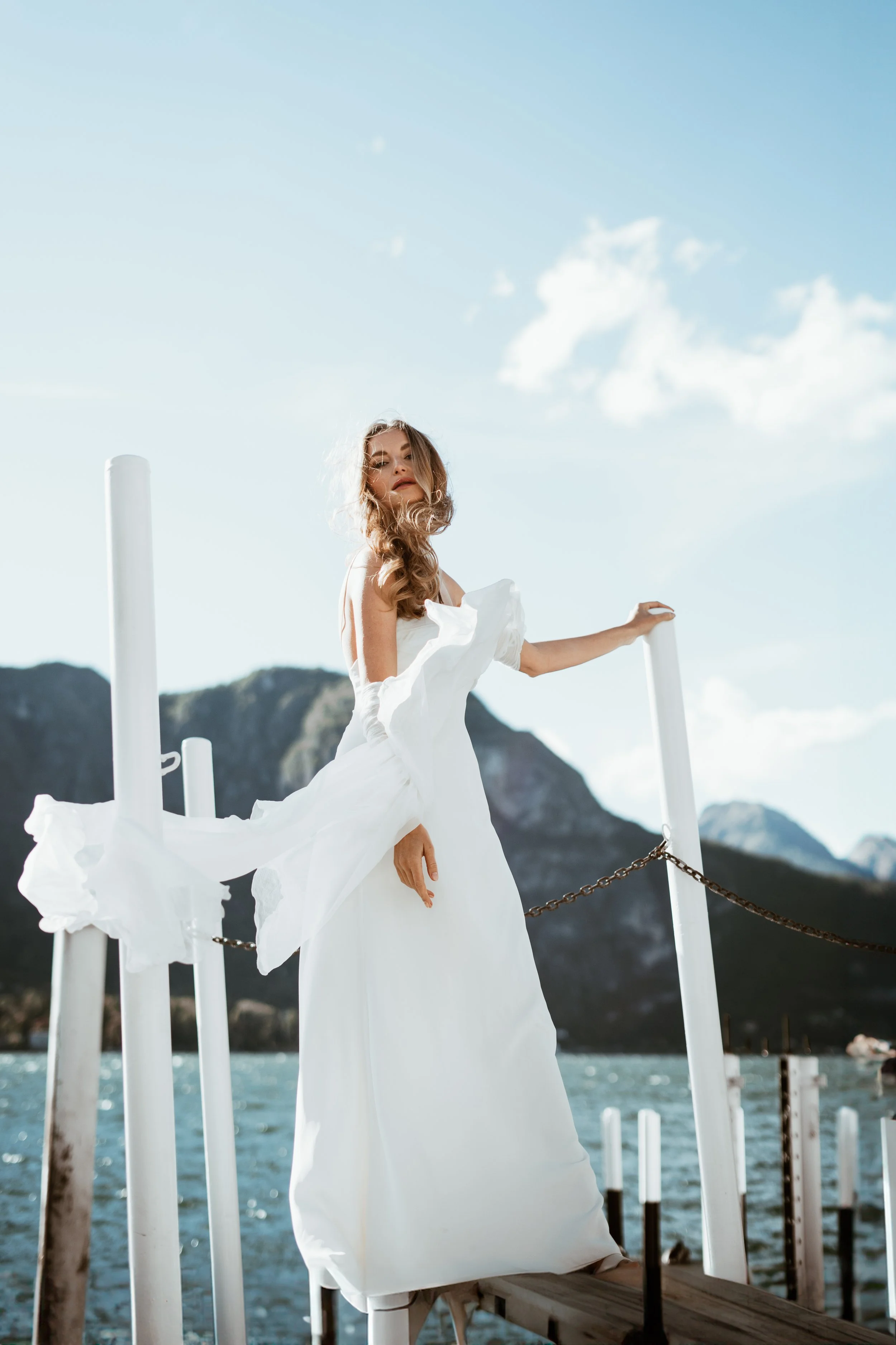 A woman in a flowing white dress standing on a dock by a body of water with mountainous terrain in the background, under a partly cloudy sky.