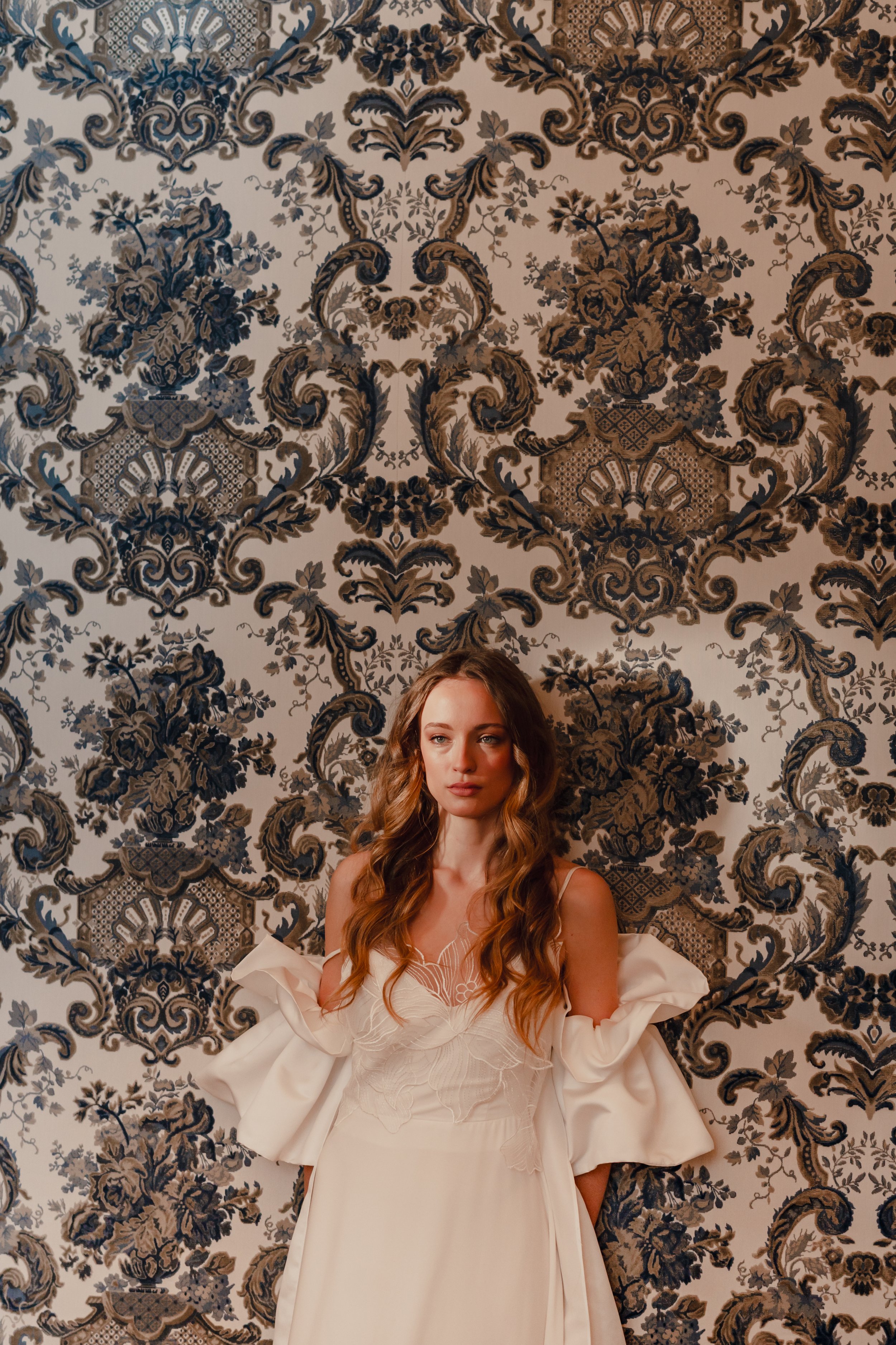 A woman with long wavy hair in a cream-colored dress with ruffled sleeves standing against an ornate, patterned wallpaper background.