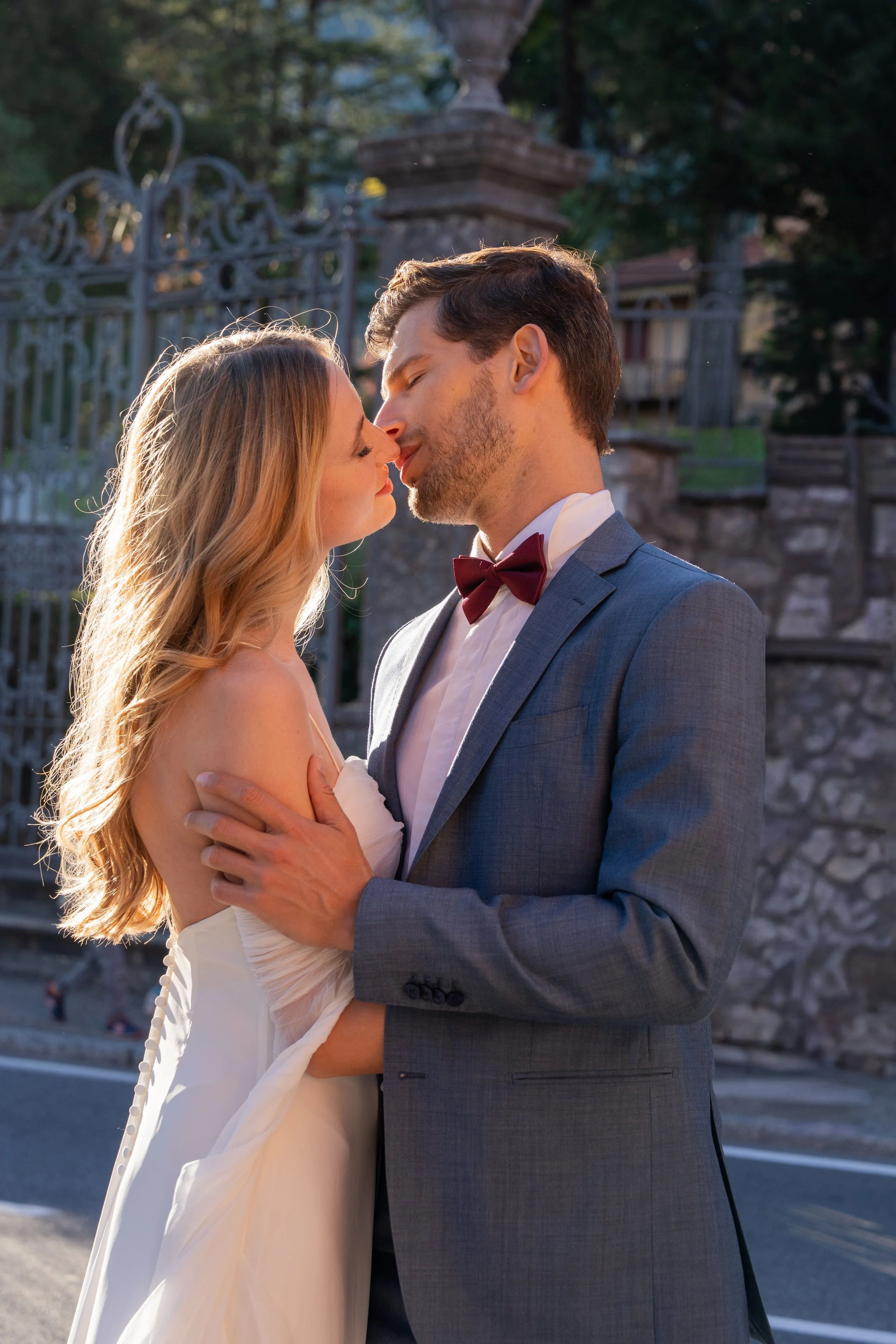 A couple in wedding attire sharing a kiss outdoors near a stone wall and decorative iron fence at sunset.