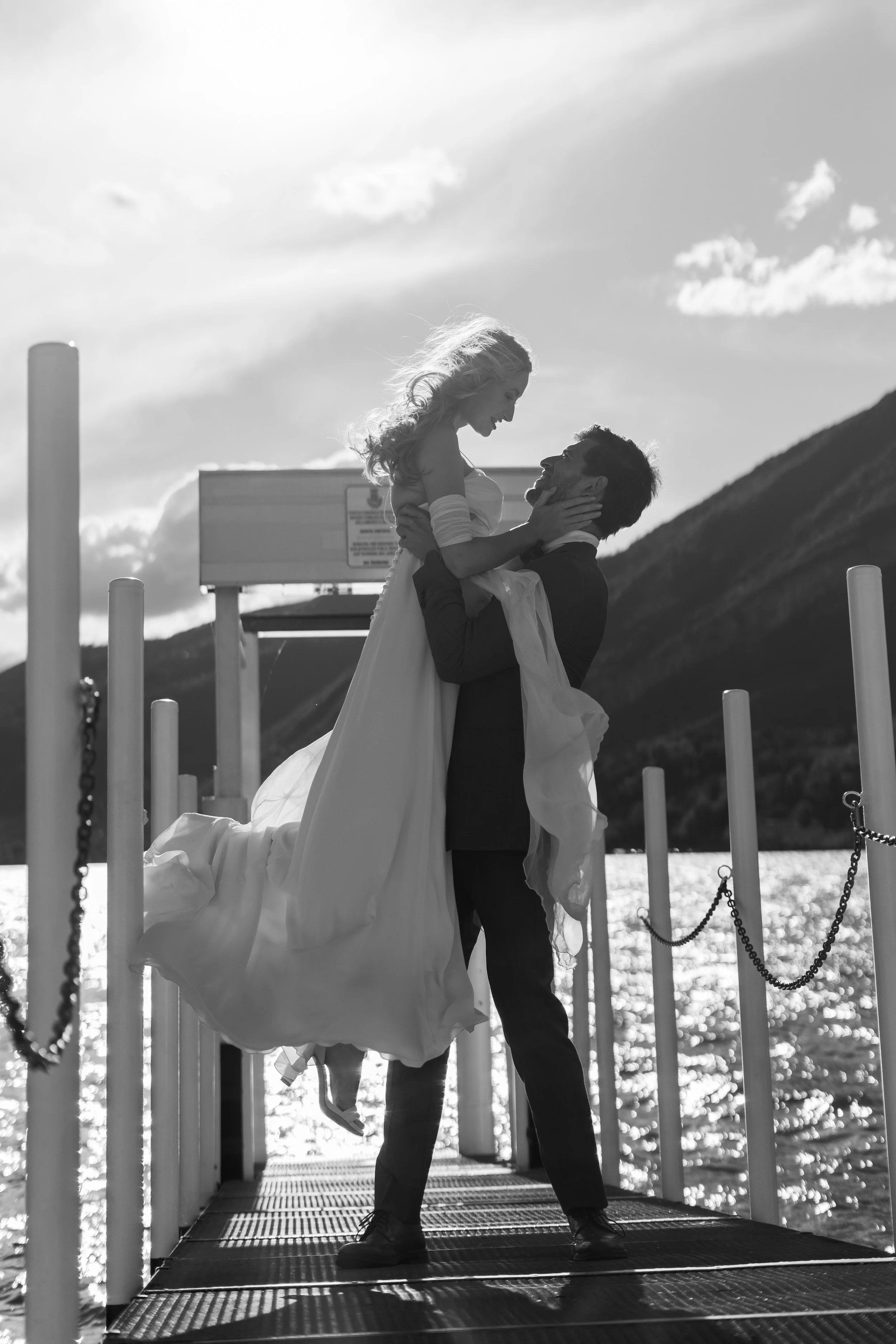 A silhouette of a couple on a dock by the water, with the man lifting the woman, both smiling, during sunset or sunrise.