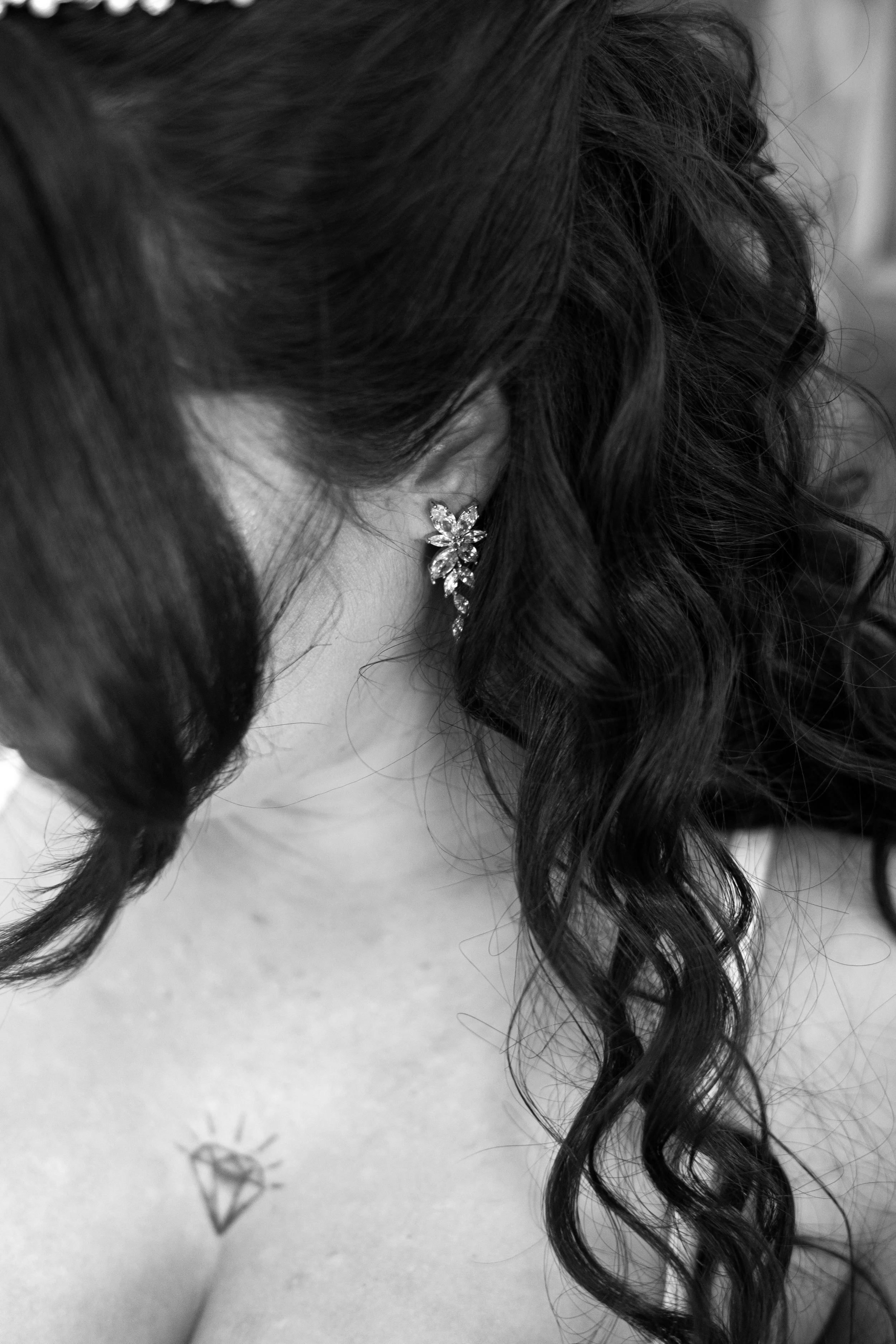 Close-up of a woman's neck and shoulder, showing an earring and a small tattoo of a diamond on her collarbone. She has dark, curly hair.