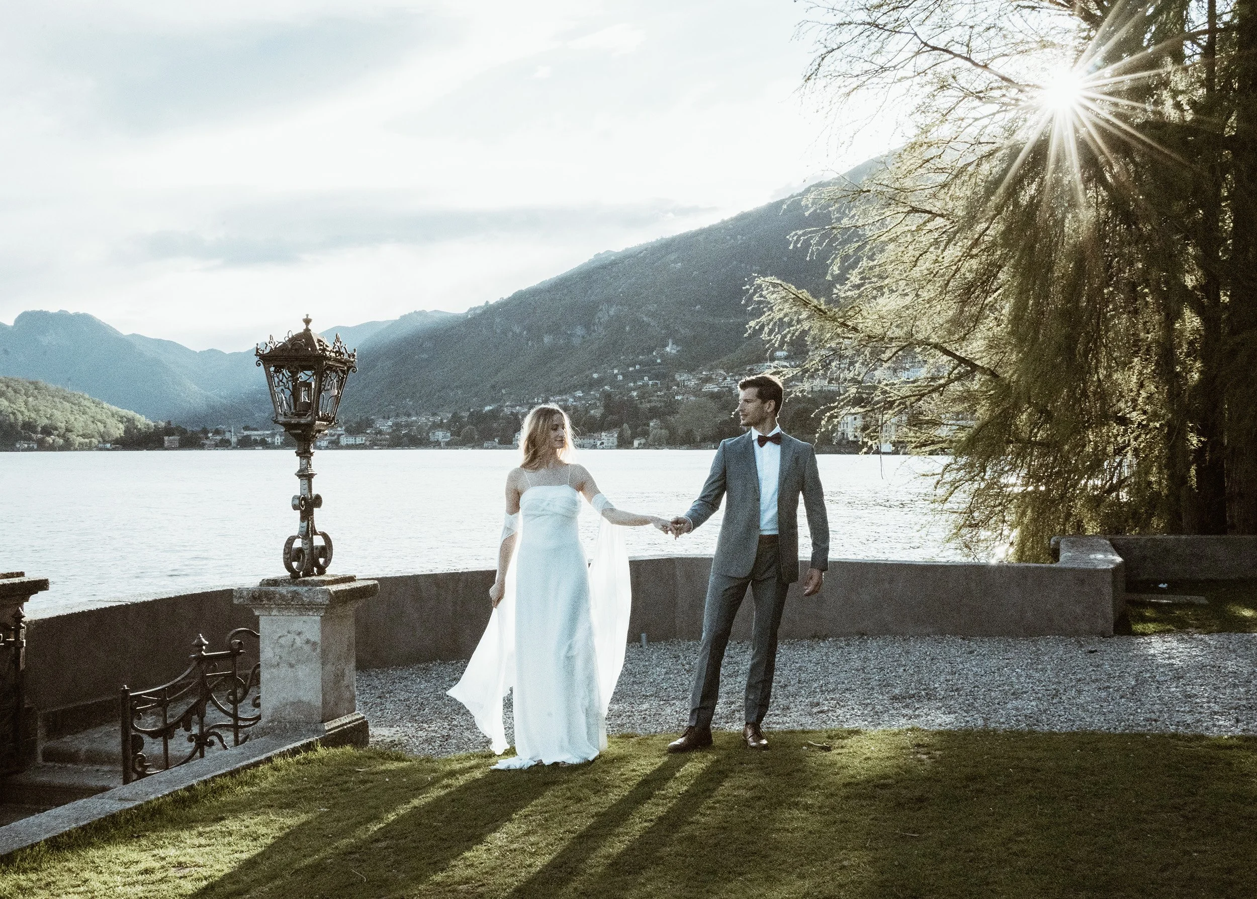 A bride and groom holding hands by a lake at sunset, with mountains and trees in the background.