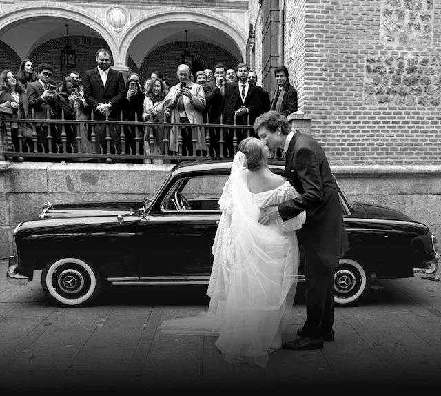 A newlywed couple kissing in front of a vintage black car during their wedding celebration, with guests watching from a balcony above.