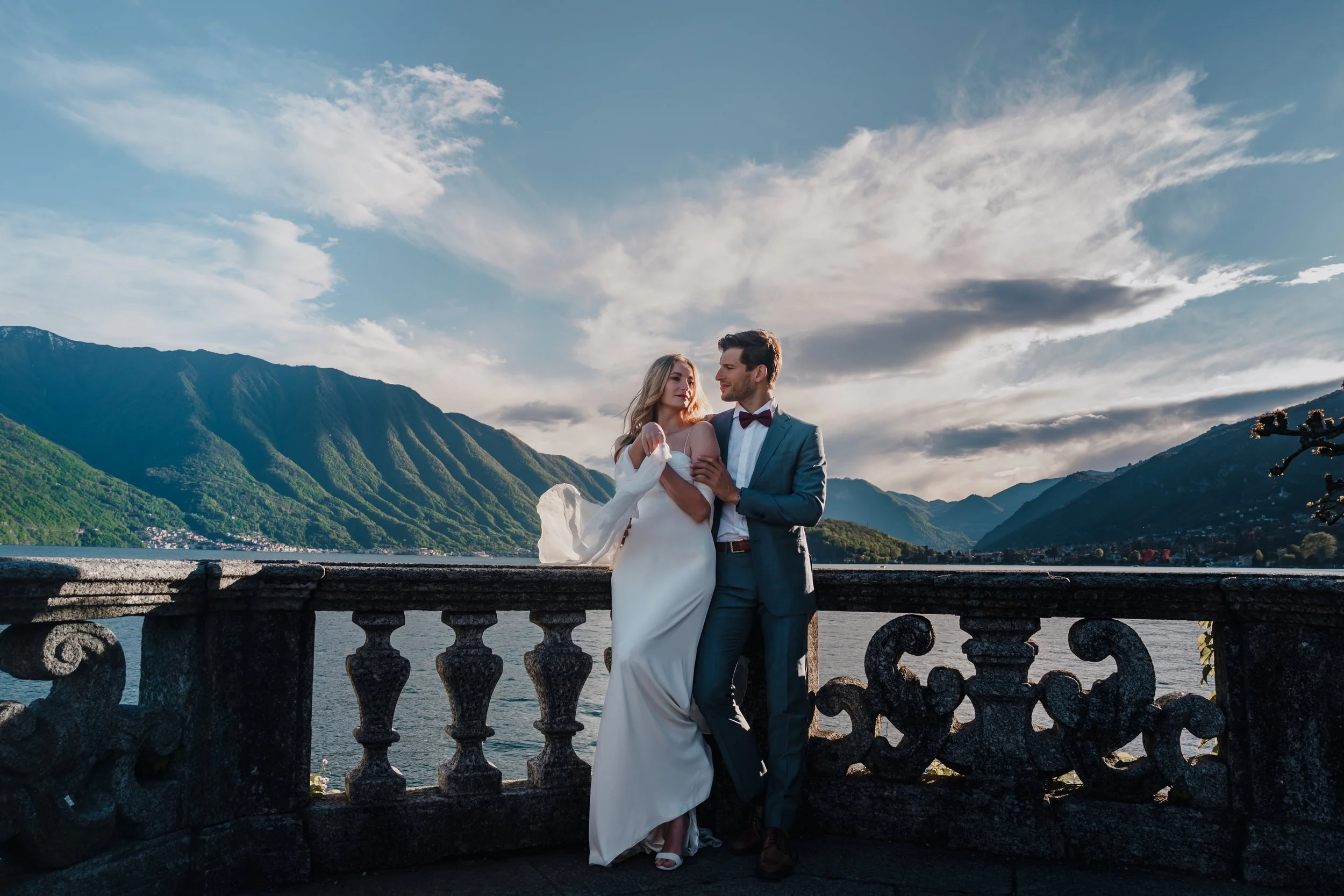 A bride and groom standing on a stone balcony overlooking a lake, surrounded by mountains under a partly cloudy sky.