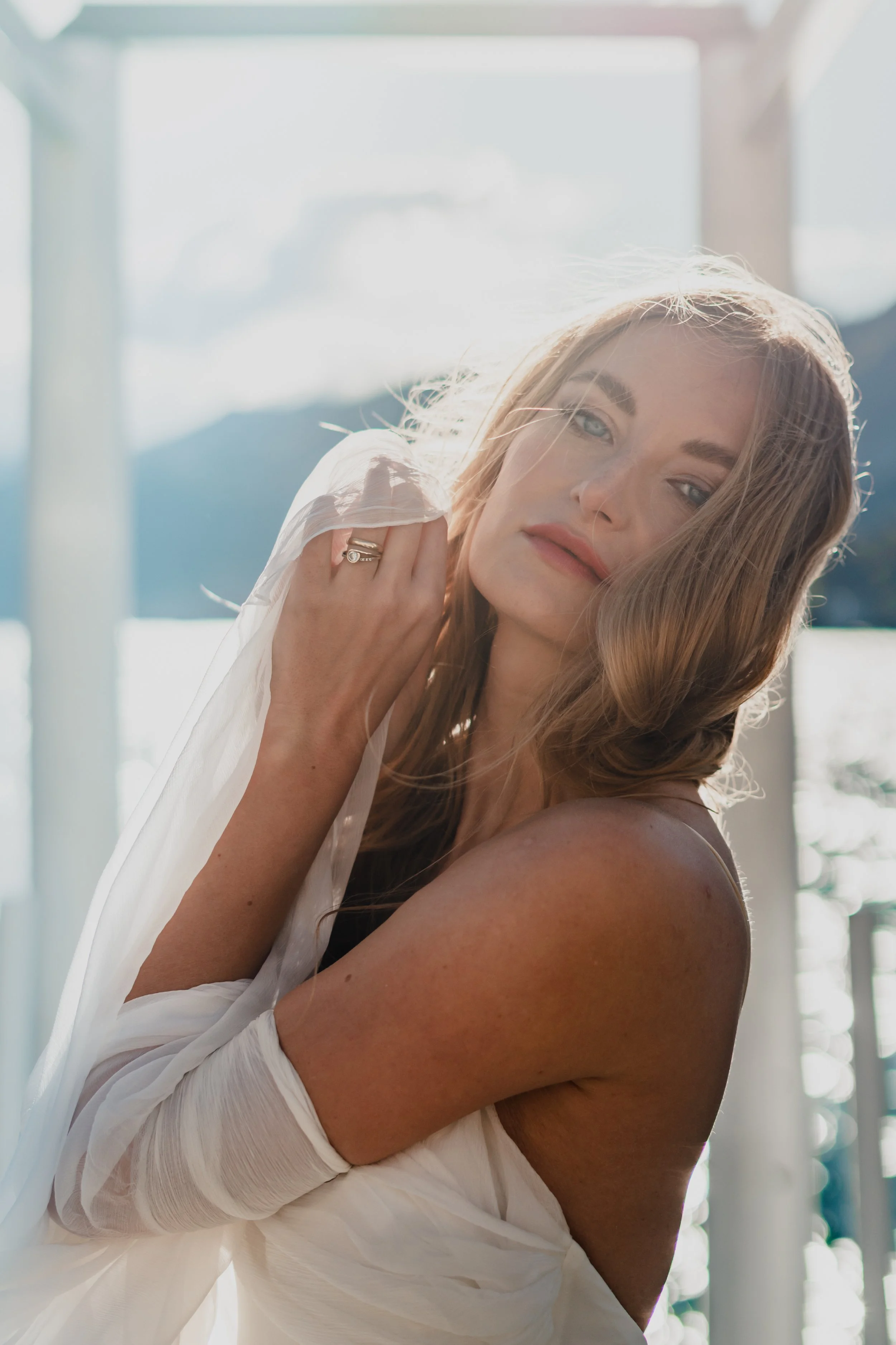 A woman with long, wavy blonde hair standing outdoors near water, wearing a white off-the-shoulder dress and a ring, with sunlight illuminating her face and hair.