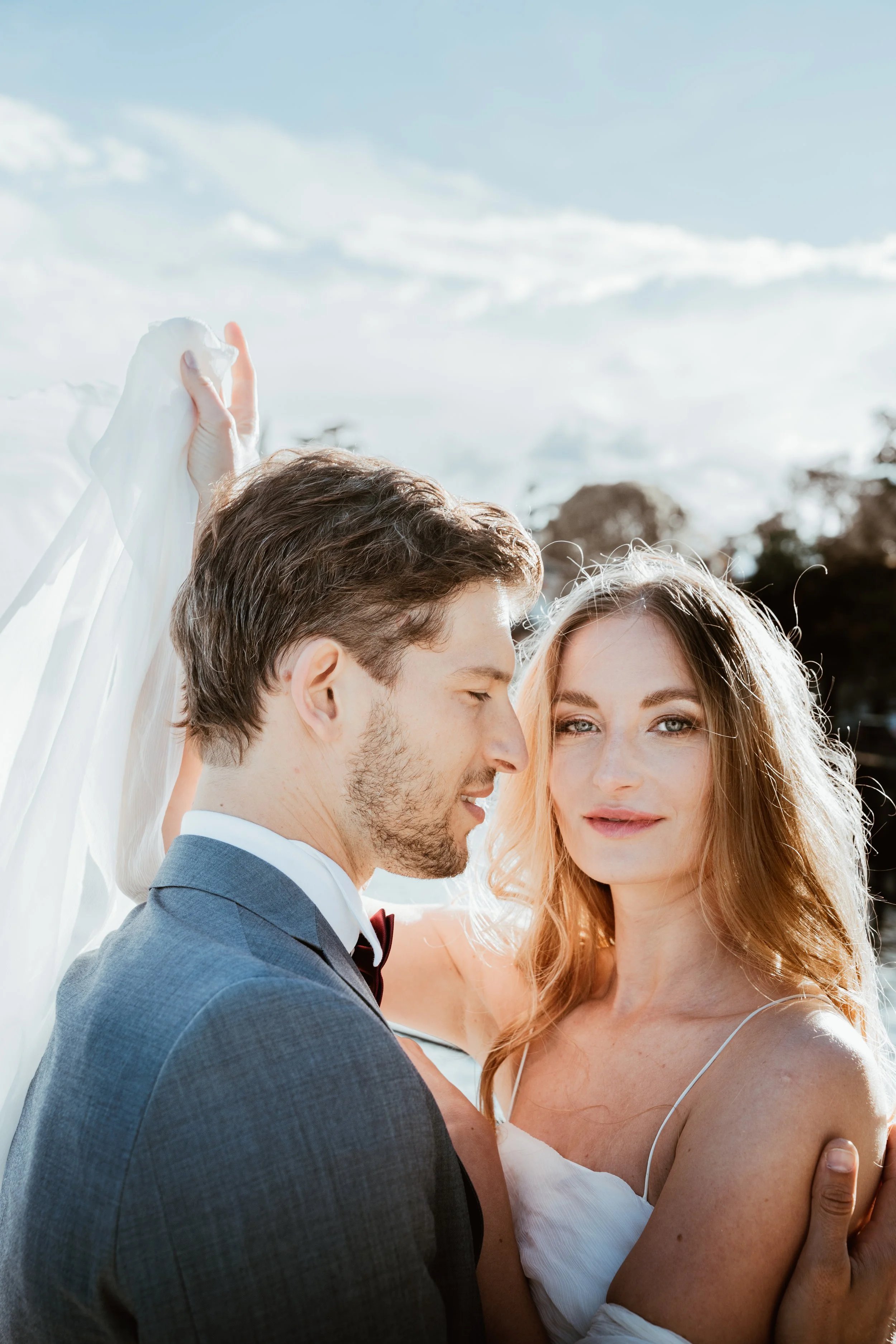 A bride and groom embracing outdoors under a bright sky, with the bride holding her veil.