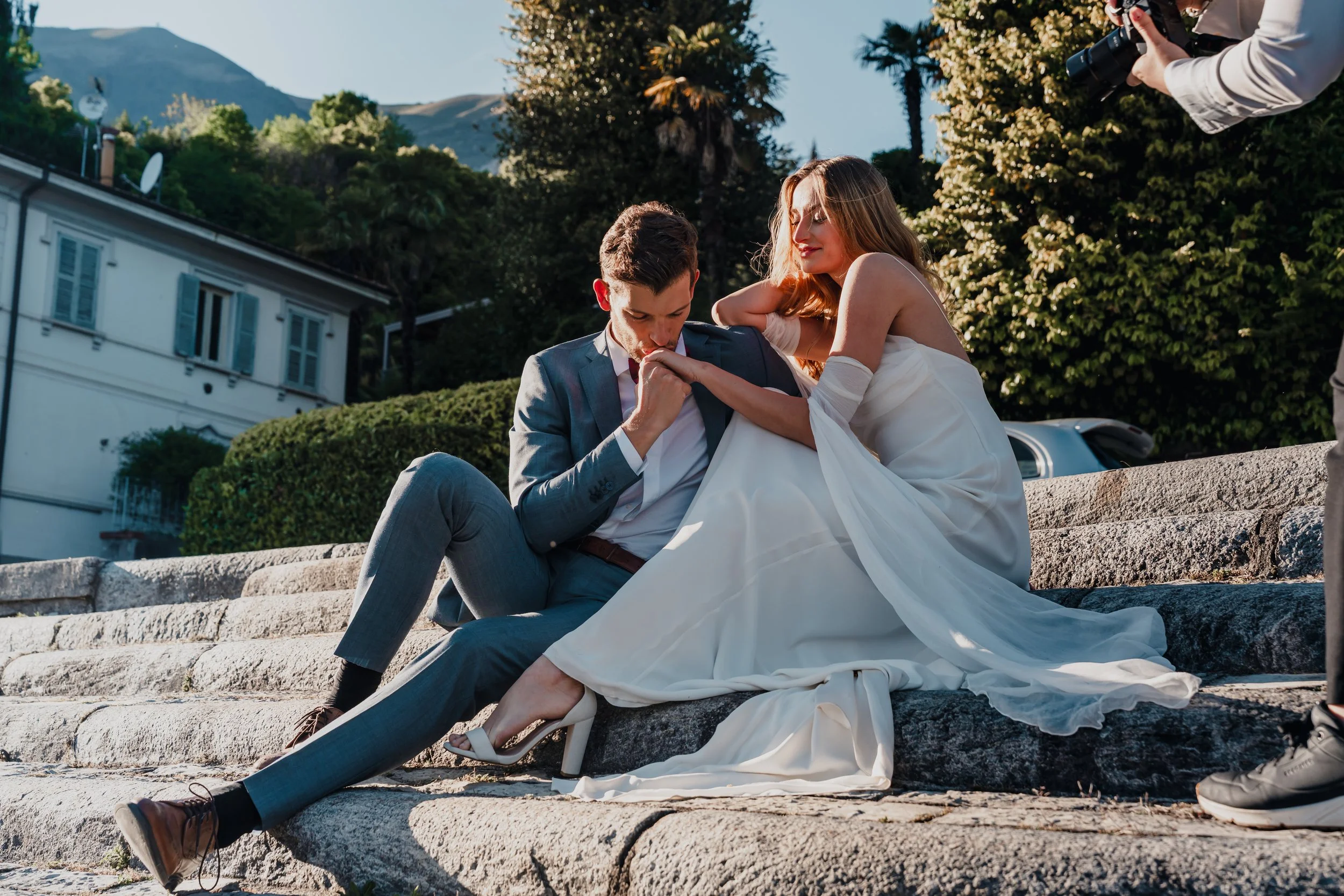 A bride and groom sit on stone steps outdoors, with the groom in a suit and the bride in a white wedding dress. The groom appears to be kissing or gently biting the bride's hand, while the bride leans toward him with a relaxed expression. A photograp