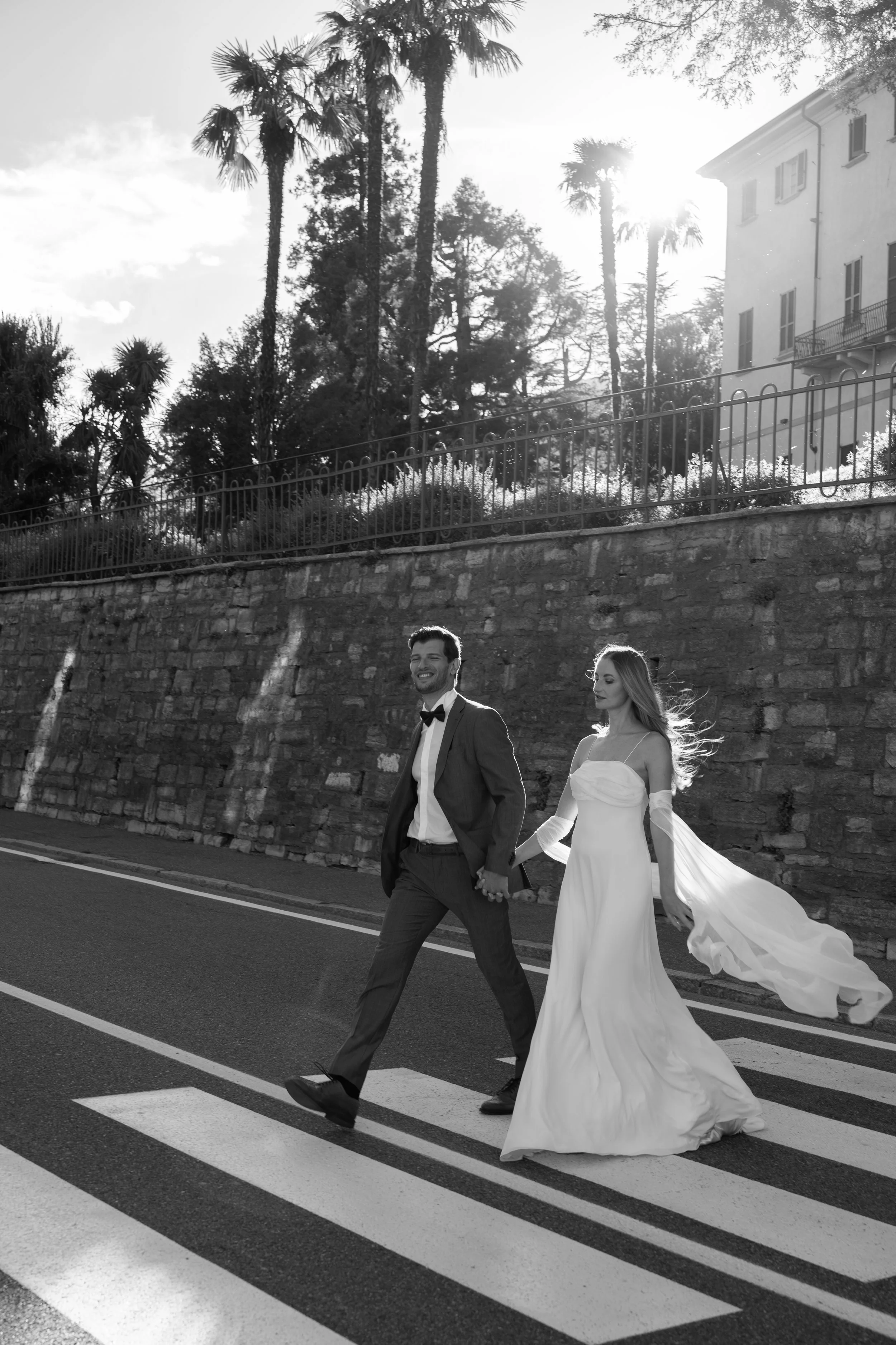 A black and white photo of a bride and groom walking hand in hand across a crosswalk, with palm trees and a building in the background.