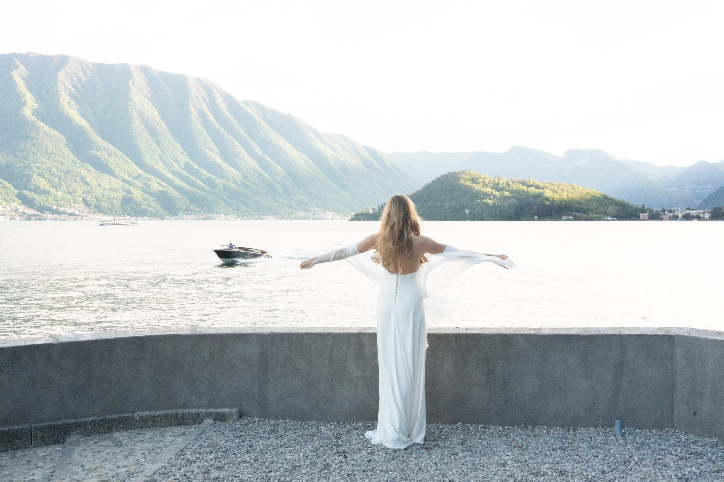 A woman in a white dress with long sleeves stands on a concrete ledge by a body of water, with arms outstretched. A boat speeds across the water, and mountains with green foliage rise in the background under a clear sky.