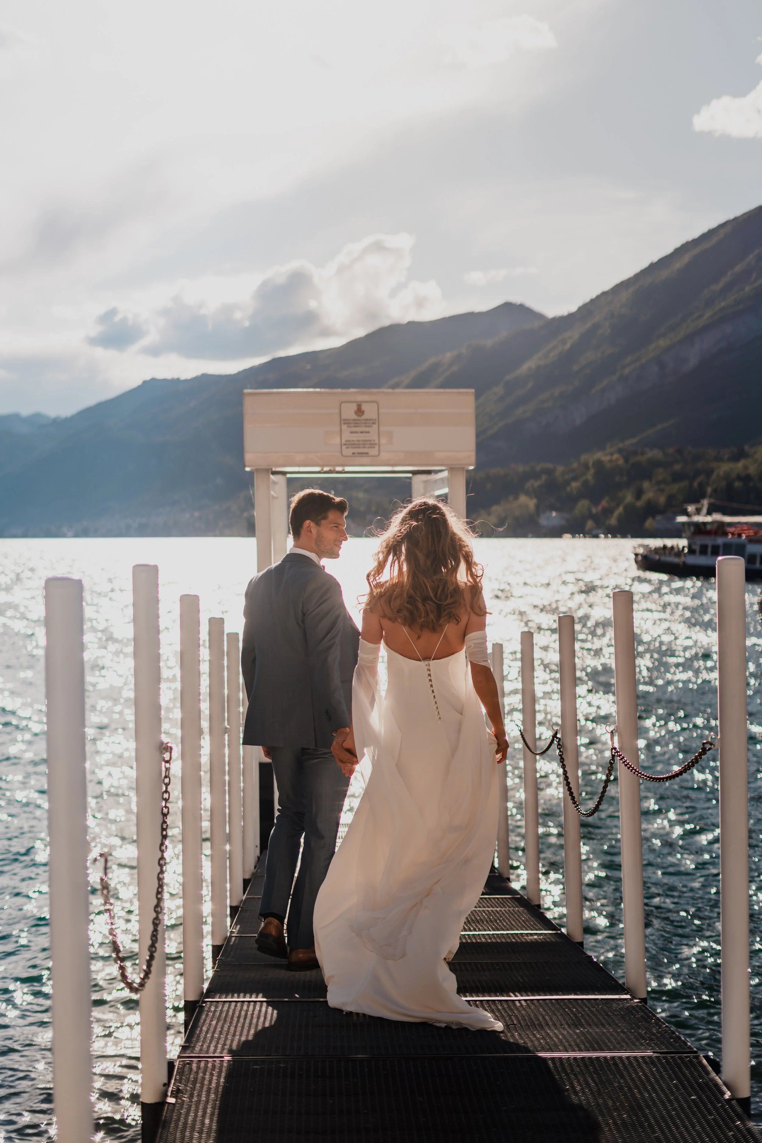 Bride and groom holding hands and walking on a dock toward a boat, with mountains and water in the background.