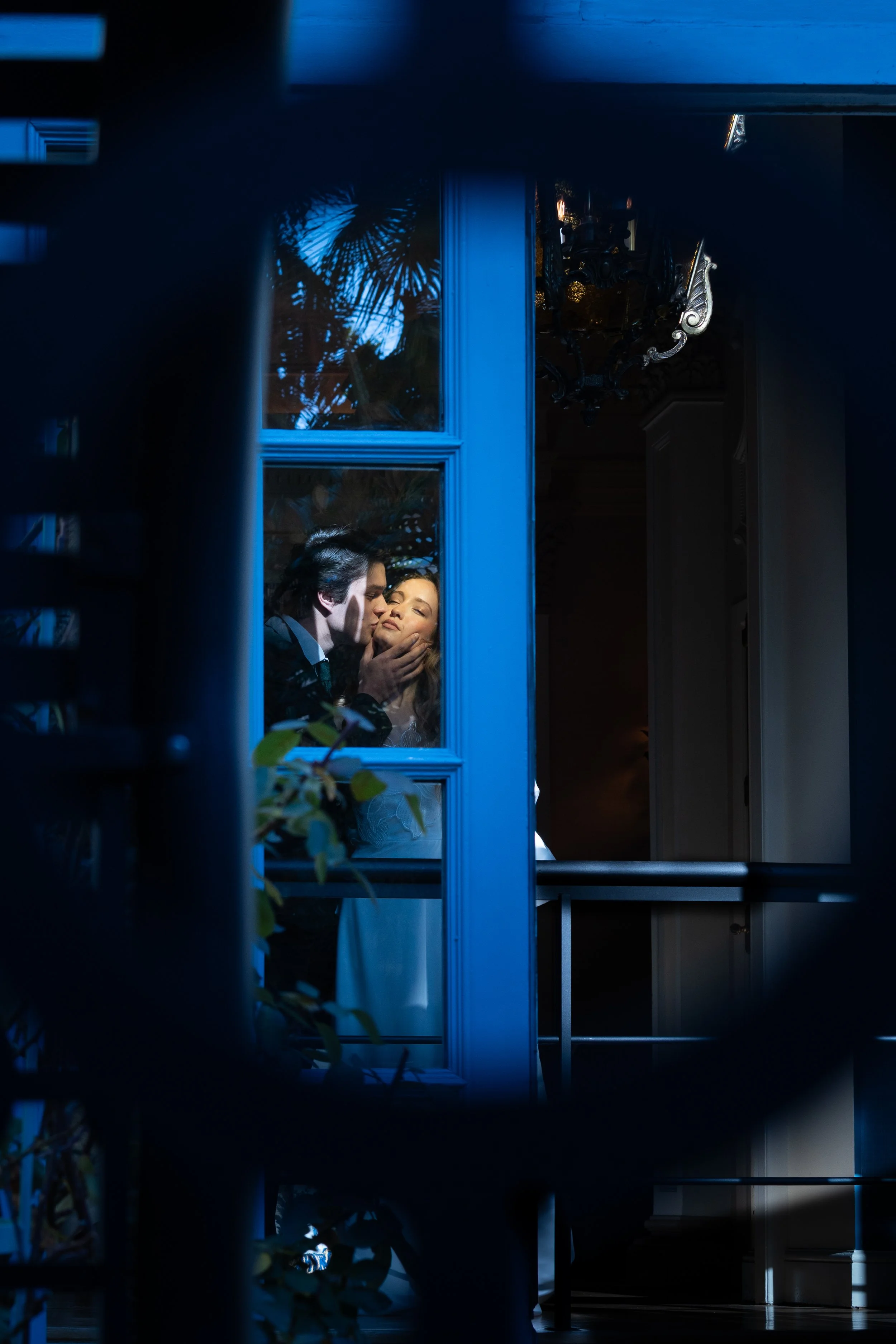 A couple sharing a romantic kiss inside a house, viewed through a window during the evening or night.