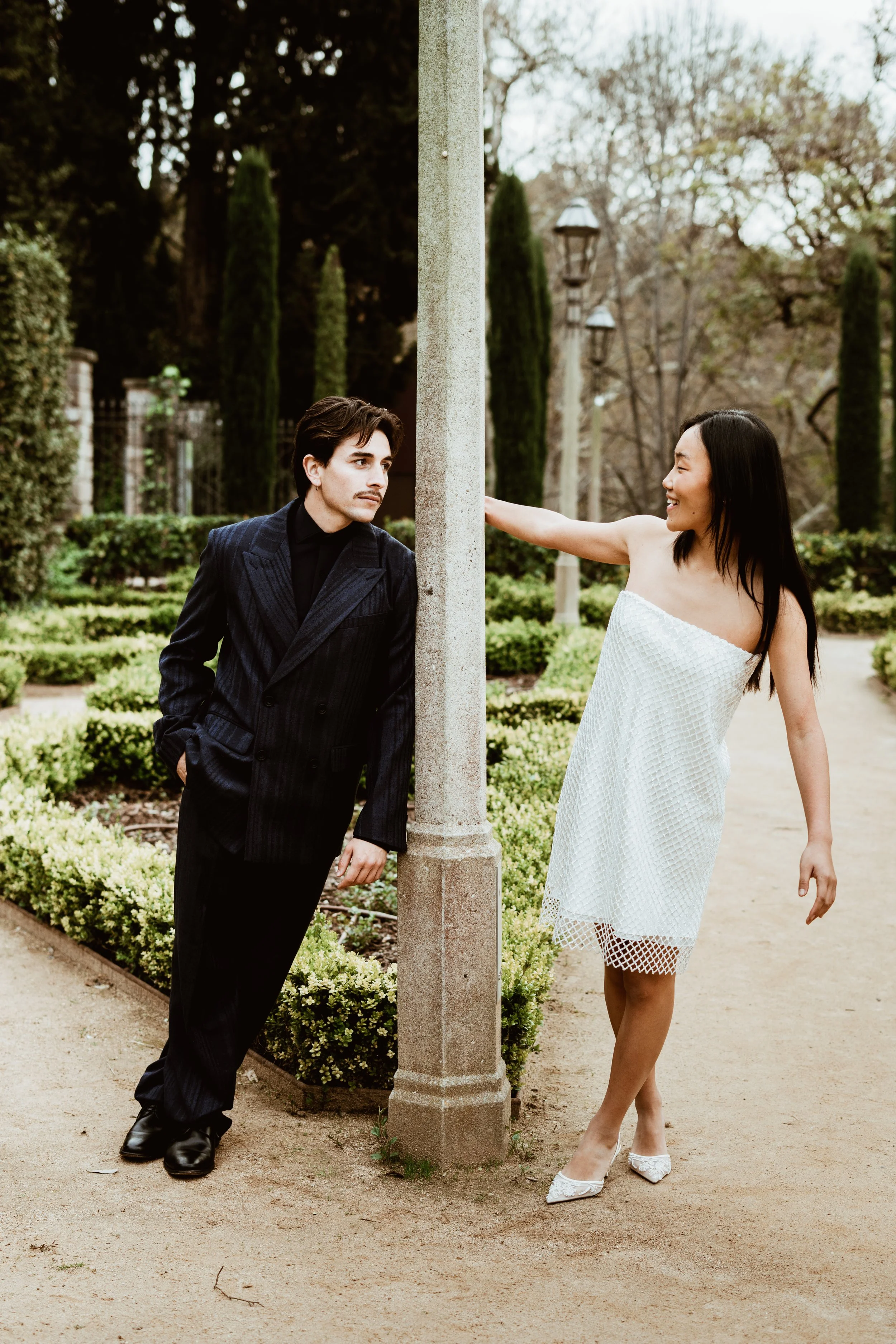 A man in a black pinstripe suit and a woman in a white strapless dress are interacting outdoors near a lamp post. The woman is smiling and touching the lamp post, while the man is leaning against it with his hands in his pockets. The background featu
