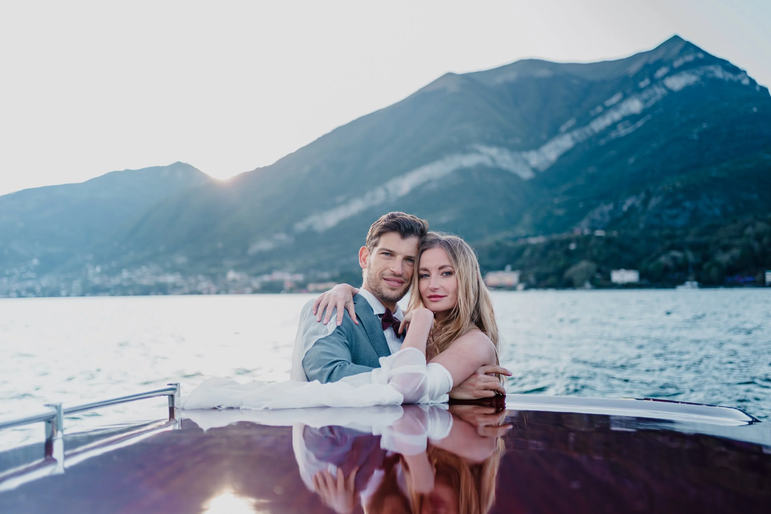 A newlywed couple in wedding attire embracing in a boat on a lake with mountains in the background during sunset.