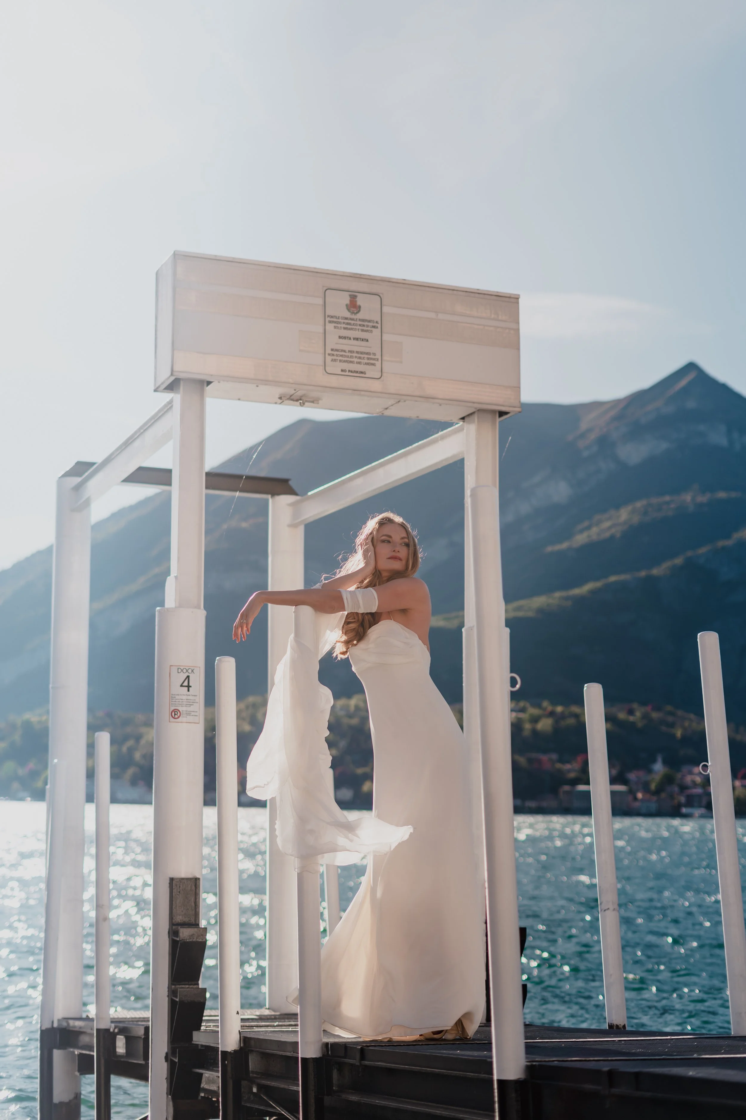 Woman in a white dress standing on a dock by a lake with mountains in the background.
