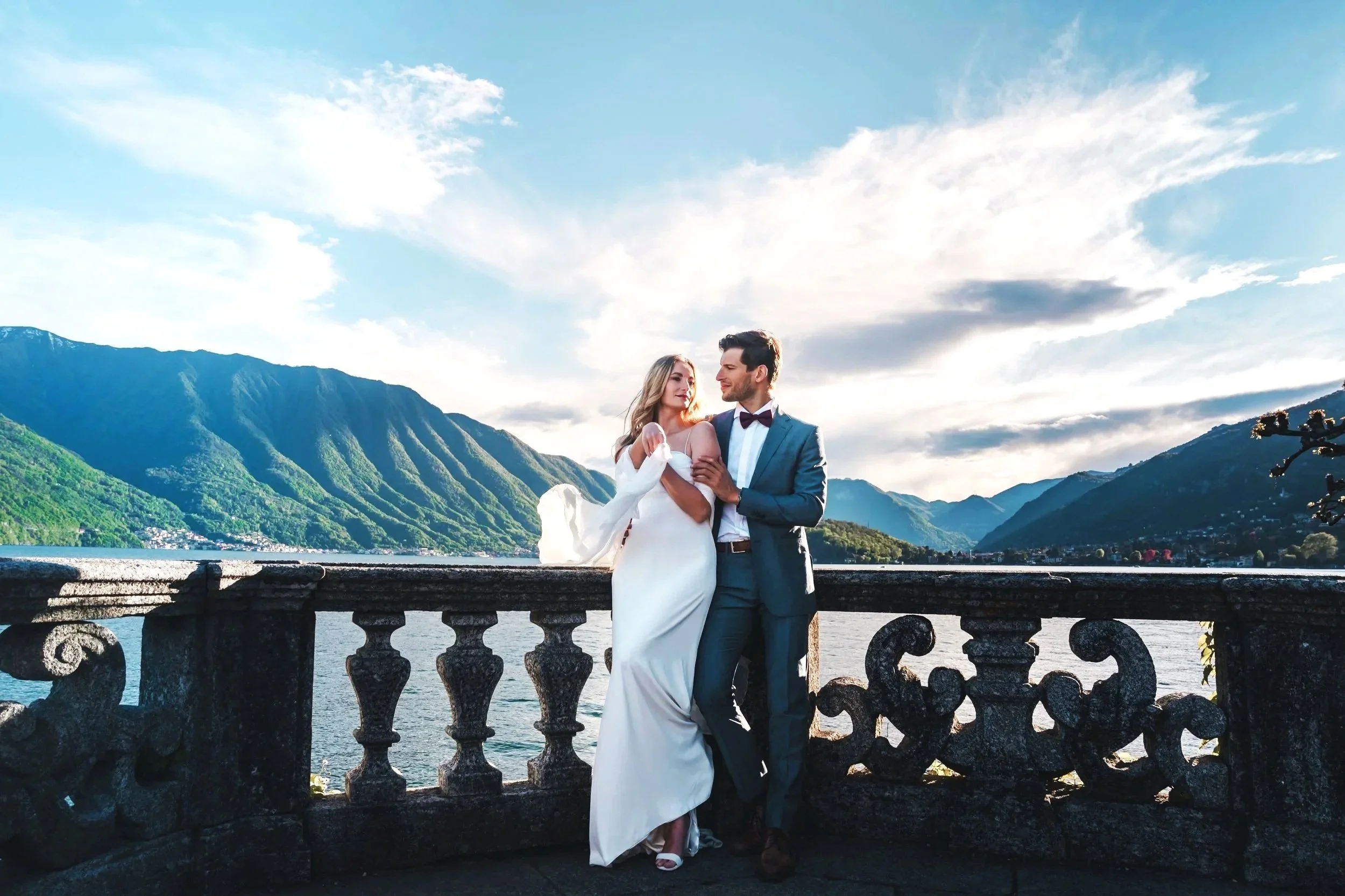 Pareja de recién casados en traje formal y vestido blanco, posando en un balcón con vista a un lago rodeado de montañas, durante el atardecer.
