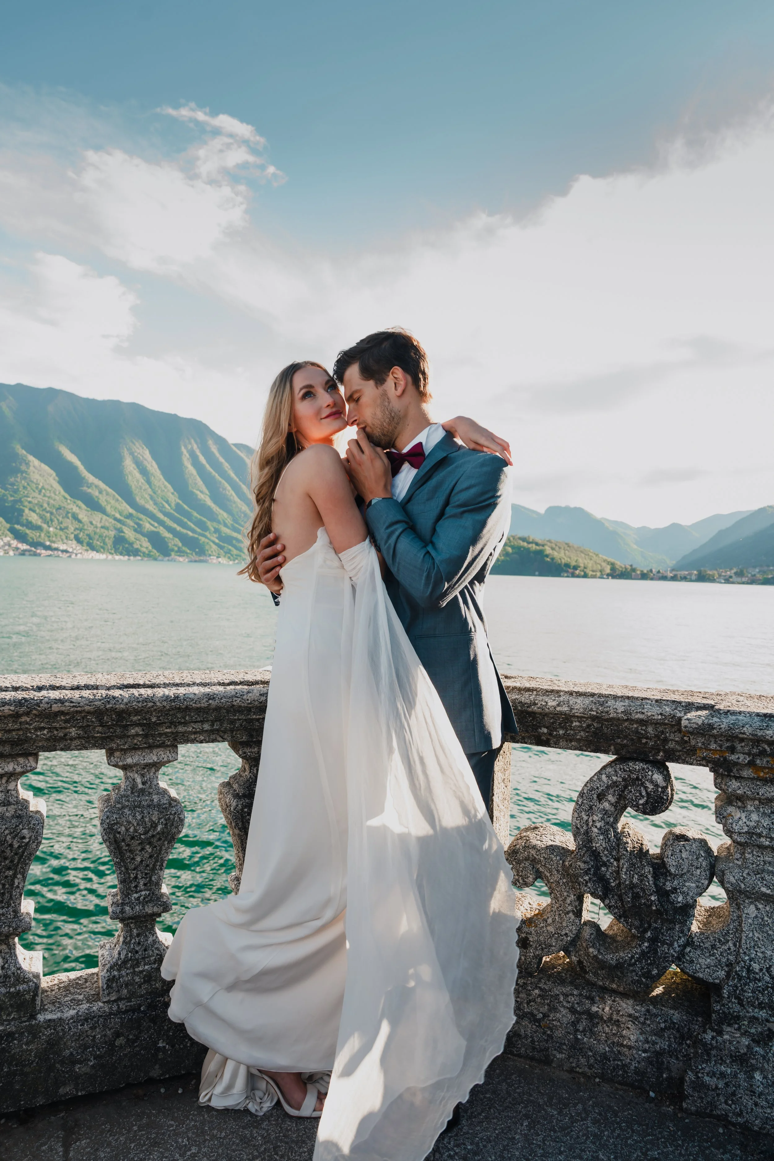 A bride and groom in wedding attire embrace on a stone balcony overlooking a lake and mountains under a partly cloudy sky.