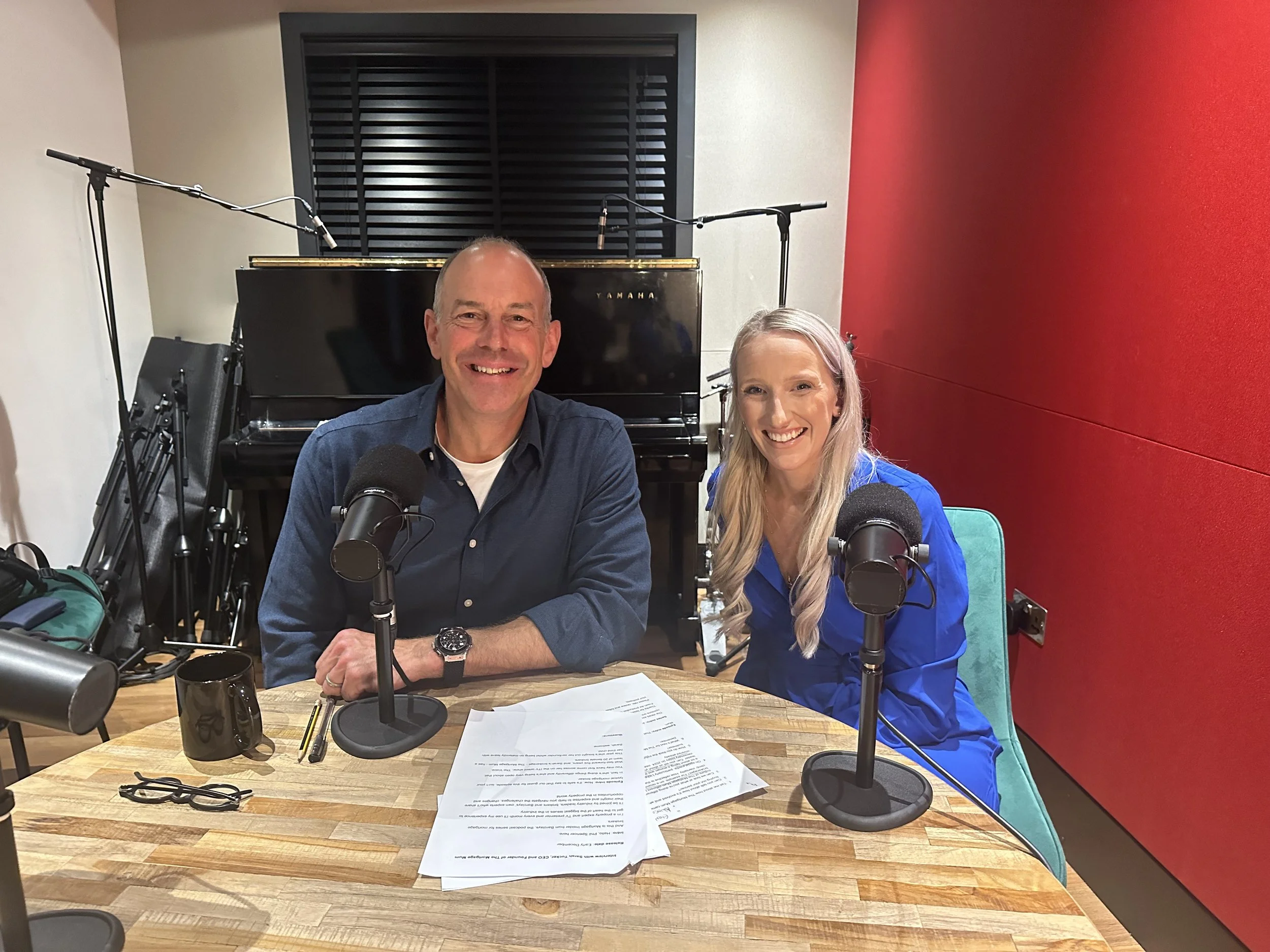 Two smiling people, a man and a woman, sitting at a table in a recording studio with microphones, a black piano in the background, and audio equipment, looking at the camera.