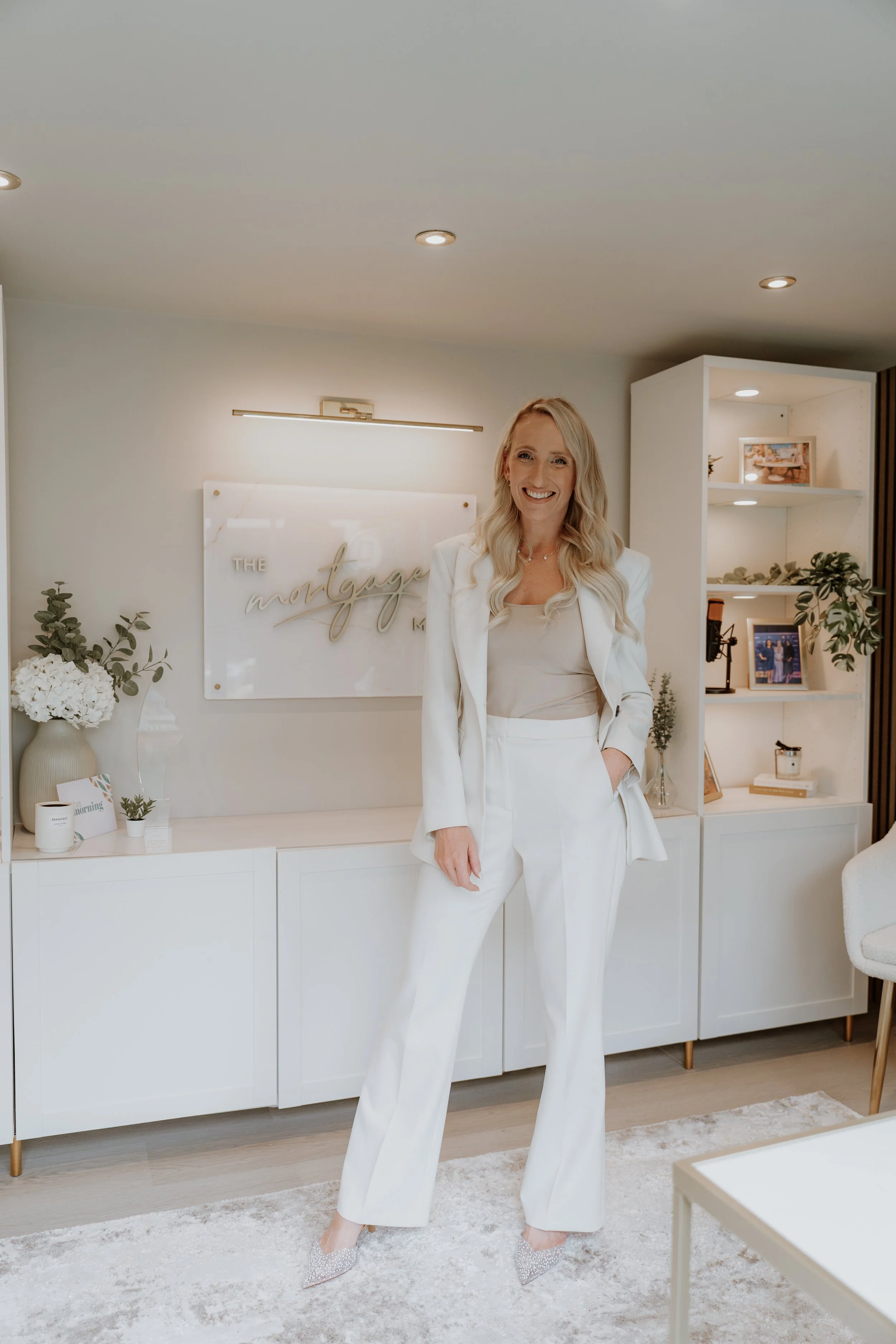 A woman dressed in a white suit standing and smiling inside a modern, well-lit office or reception area with a white cabinet, floral arrangements, and framed photos.