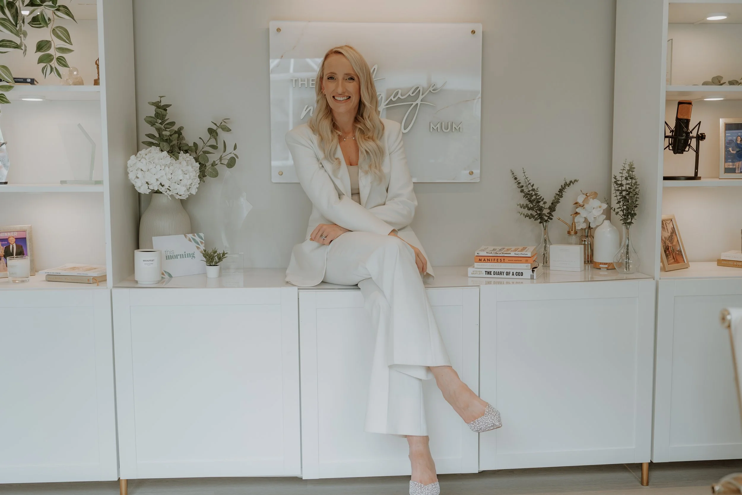 A woman with long blonde hair, dressed in a white suit, sitting on a white cabinet in a modern, minimalist room, smiling at the camera. Behind her is a sign that reads, 'The Vintage Mum,' with shelves containing books, framed pictures, and decorative items, and various vases with white flowers and greenery.