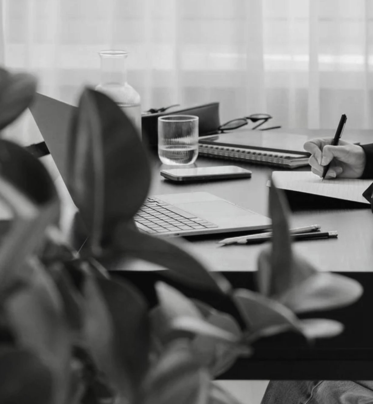 A black-and-white photo of a workspace with a hand taking notes, glasses, a notebook, a tablet, a glass of water, and office supplies on a table.