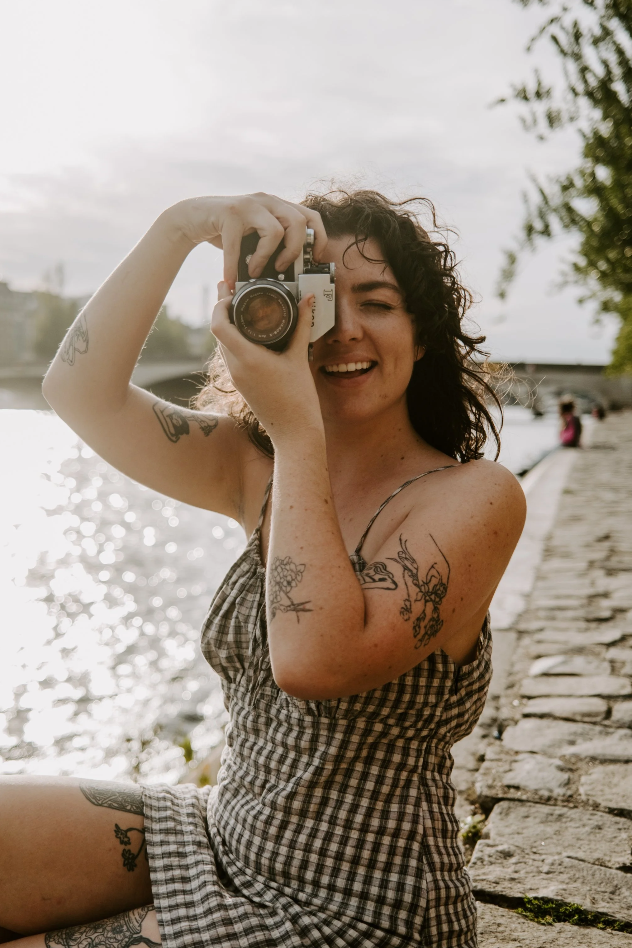 A woman with curly dark hair taking a photo with a camera by a waterfront, sitting on a cobblestone path, smiling, wearing a checkered dress, with tattoos on her arms and thigh.