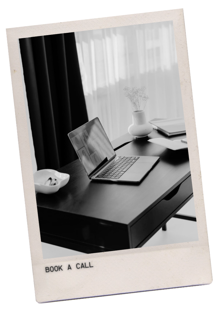 A Polaroid-style black and white photo of a tidy desk with a laptop, a white vase with a plant, open notebooks, and a small dish, with curtains and a window in the background.