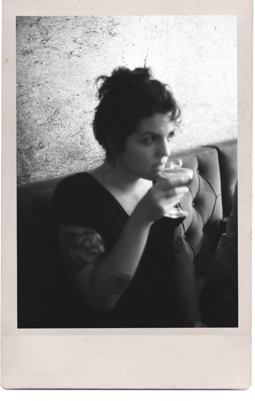 A woman with short curly hair sitting on a cushioned seat, holding a cocktail glass with a drink, looking to her right in a dimly lit setting with a textured wall behind her.
