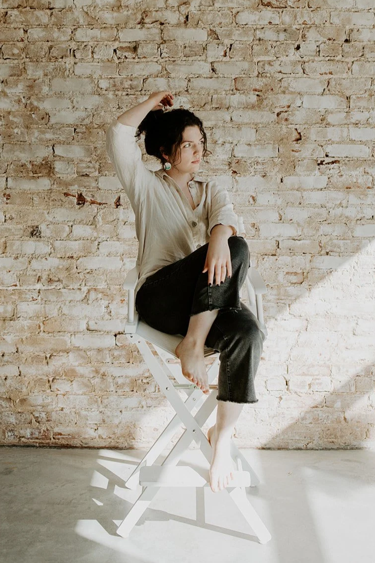 A woman with curly dark hair sitting barefoot on a white folding chair against a brick wall. She is wearing a light-colored button-up shirt and black jeans, with one hand on her head and the other resting on her knee, looking to the side.