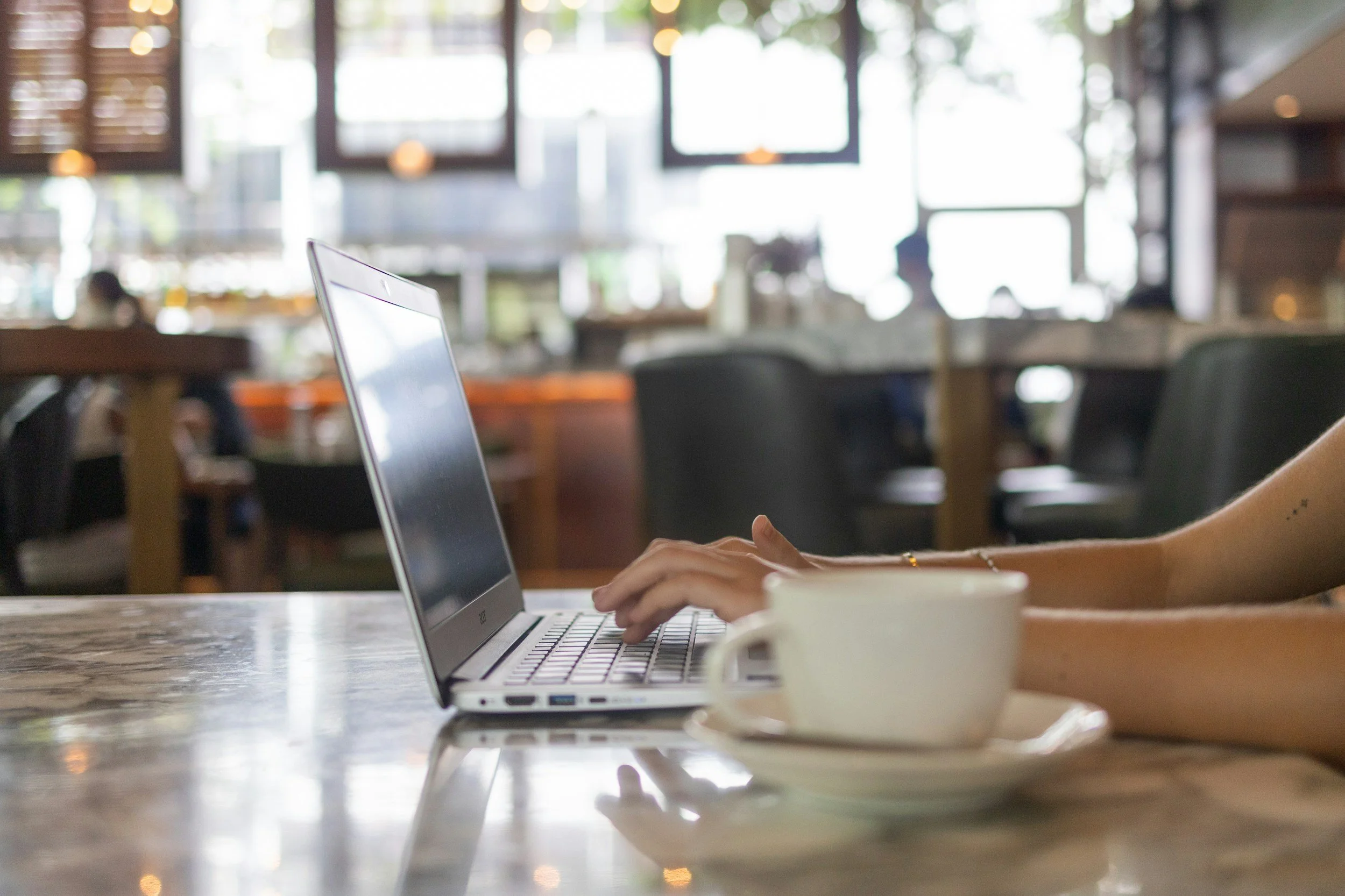 Person working on a laptop with a coffee cup on a saucer in a coffee shop.