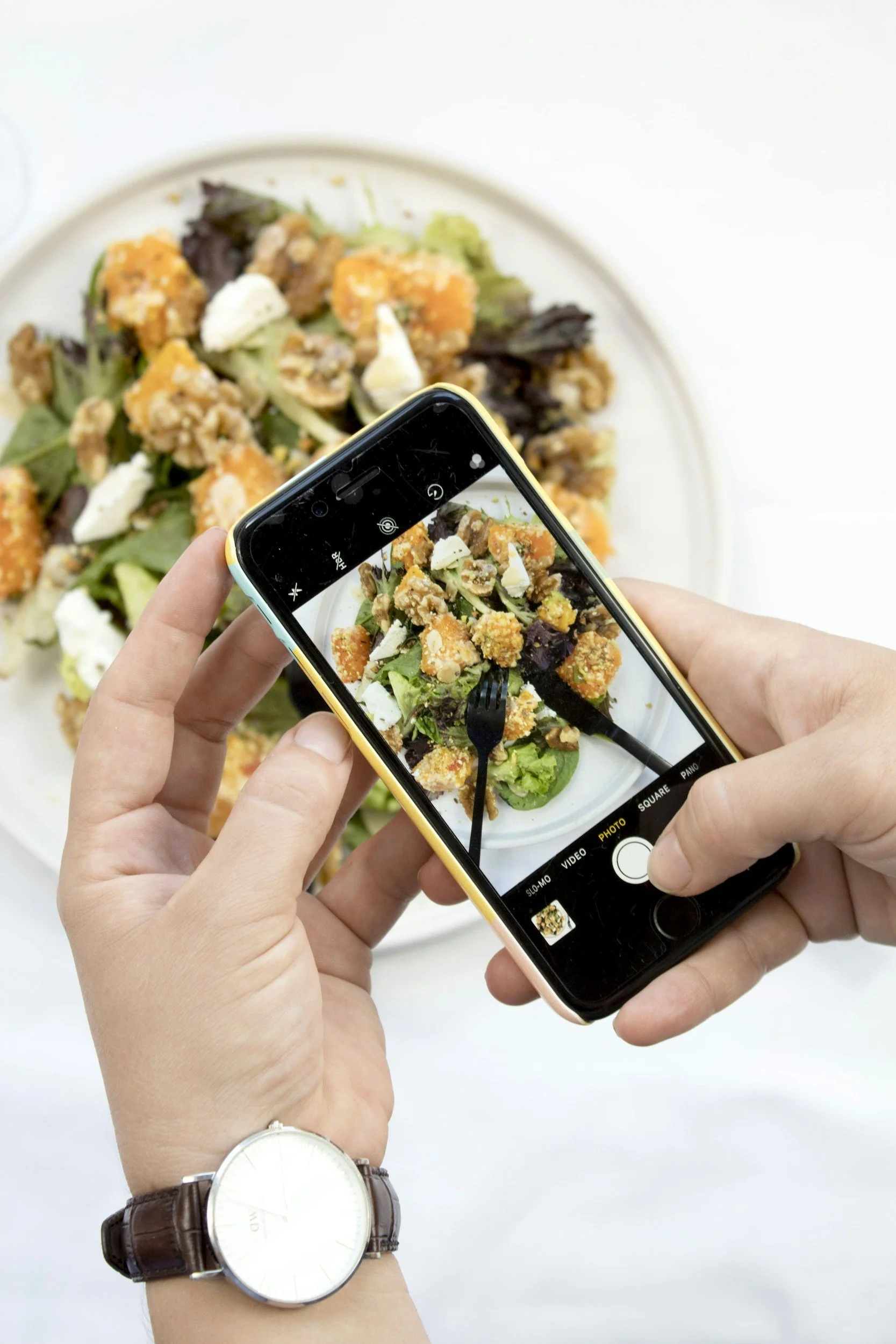 Person taking a photo of a salad on a white plate with a smartphone, showing mixed greens, croutons, and cheese.