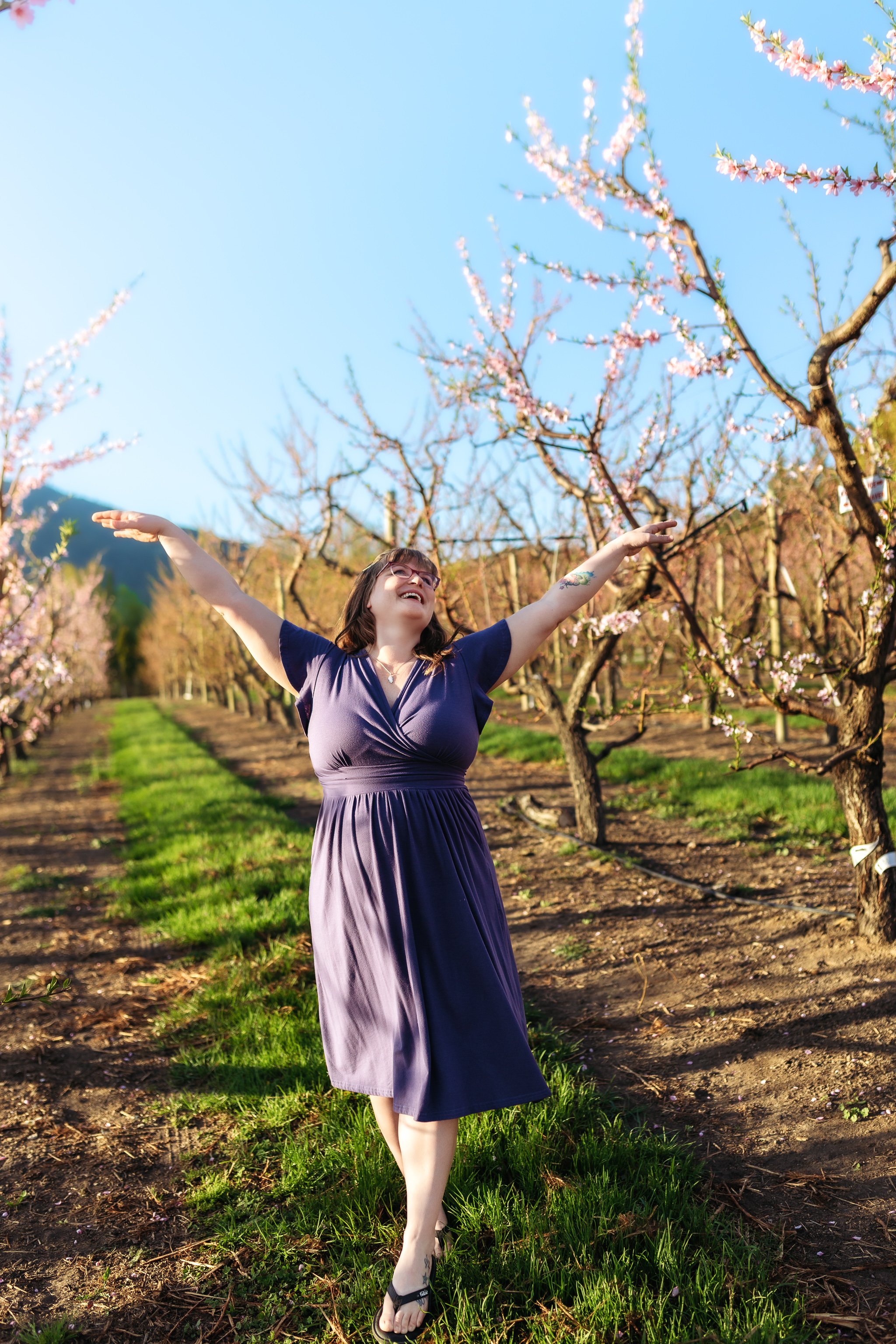 Woman in a purple dress joyfully standing with arms outstretched in an orchard with blooming peach trees during spring.