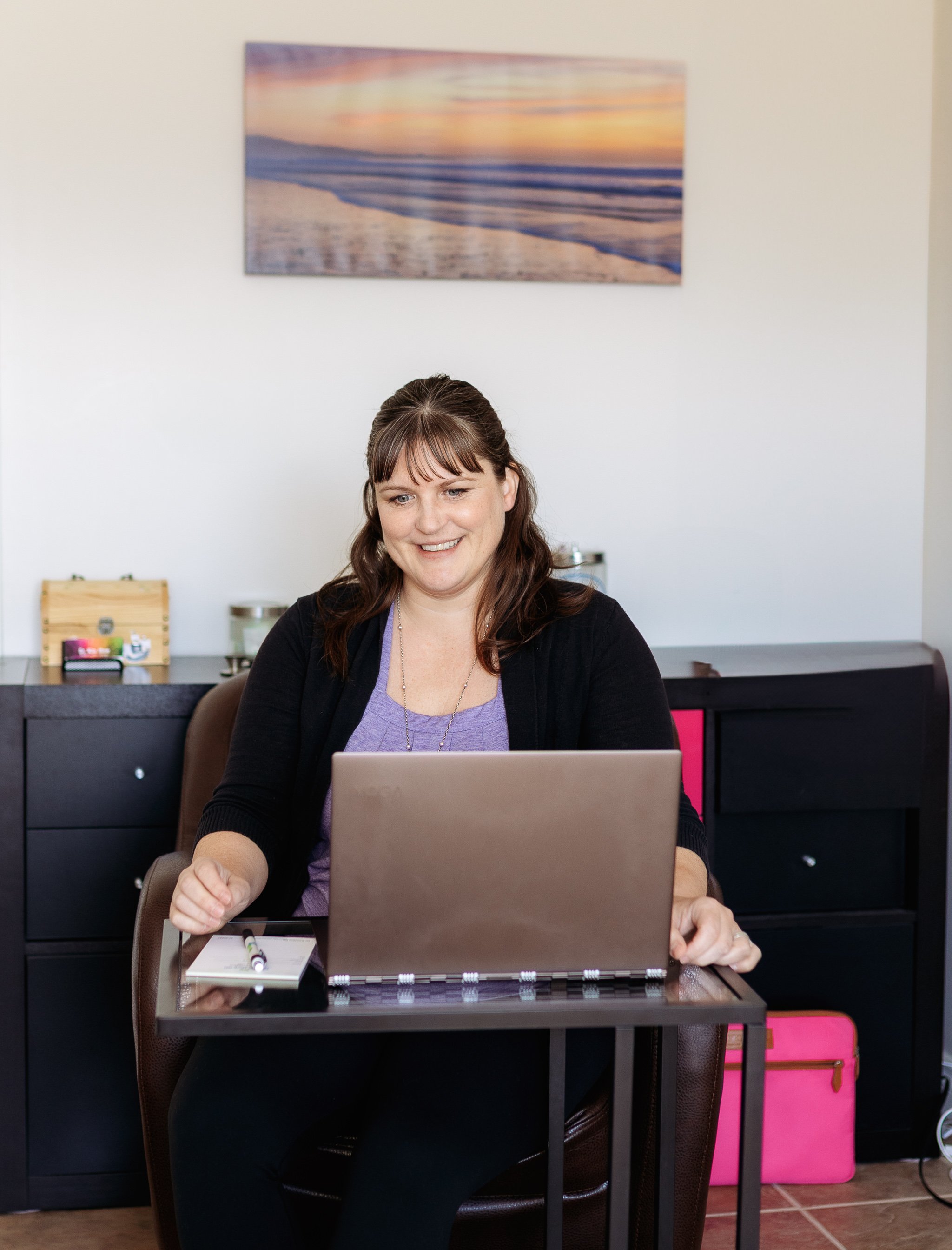 A woman sitting at a desk with a laptop, smiling, with a sunset beach scene artwork on the wall behind her.