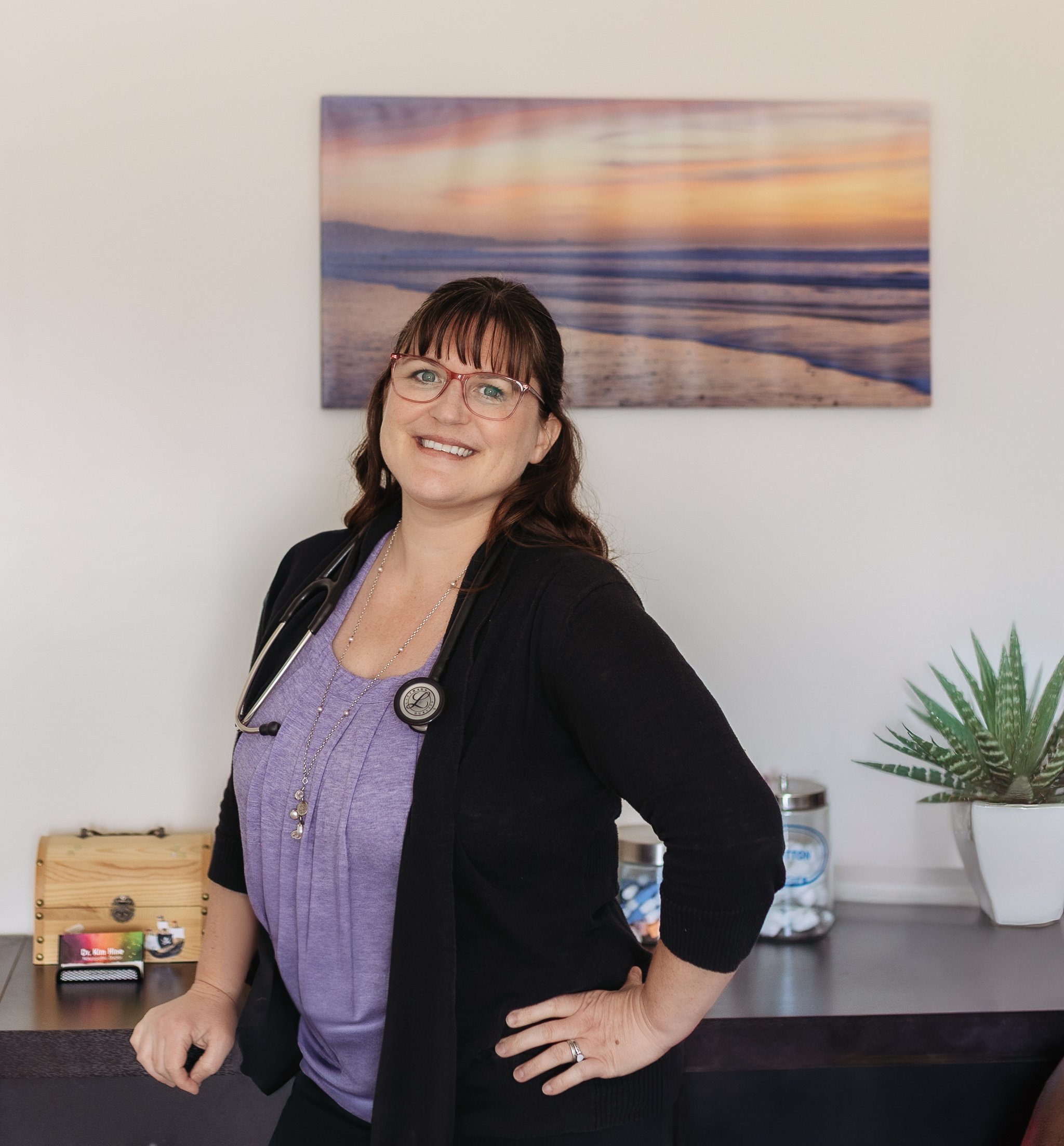 A smiling female healthcare professional wearing glasses and a stethoscope around her neck, standing indoors near a desk with medical supplies, a succulent plant, and a mountain and sunset painting in the background.
