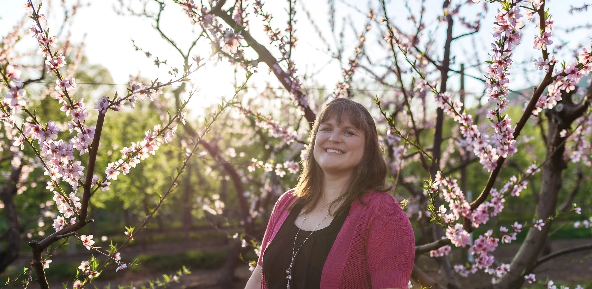 A woman smiling outdoors among blooming pink flowers on trees, with sunlight filtering through the branches in the background.
