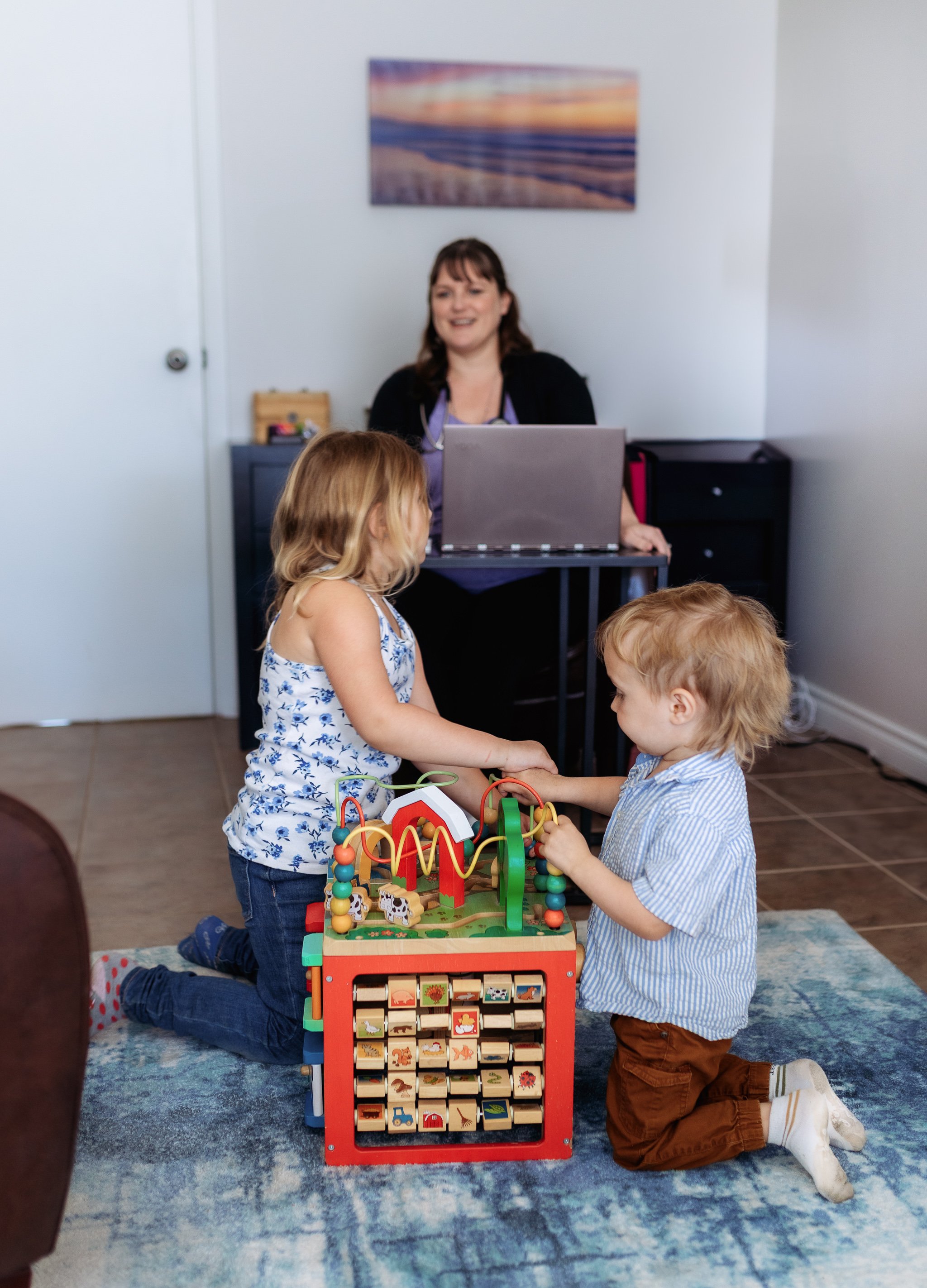 Two young children, a girl and a boy, kneel on a blue rug in a room, playing with a colorful wooden toy. A woman sits behind a desk with a laptop, watching them and smiling. The room has white walls, a painting of a beach scene, and a black cabinet.