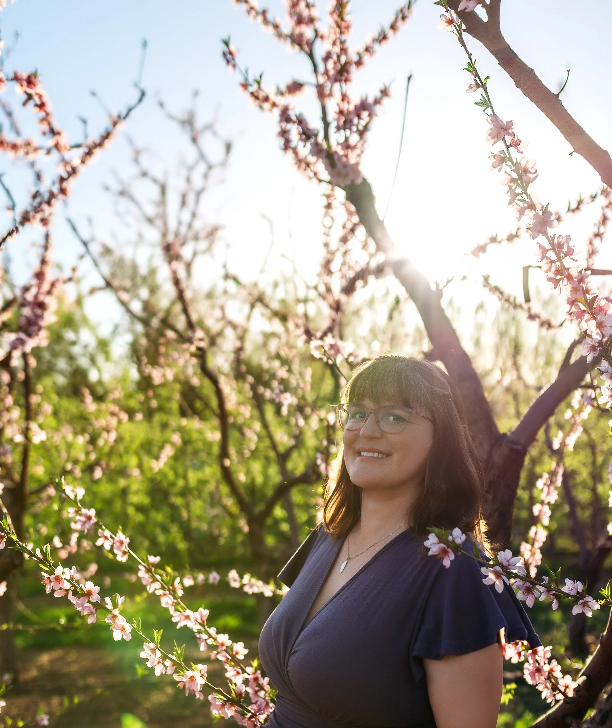 A woman with glasses and brown hair smiling outdoors in a garden of blooming pink flowers with the sun shining behind her.