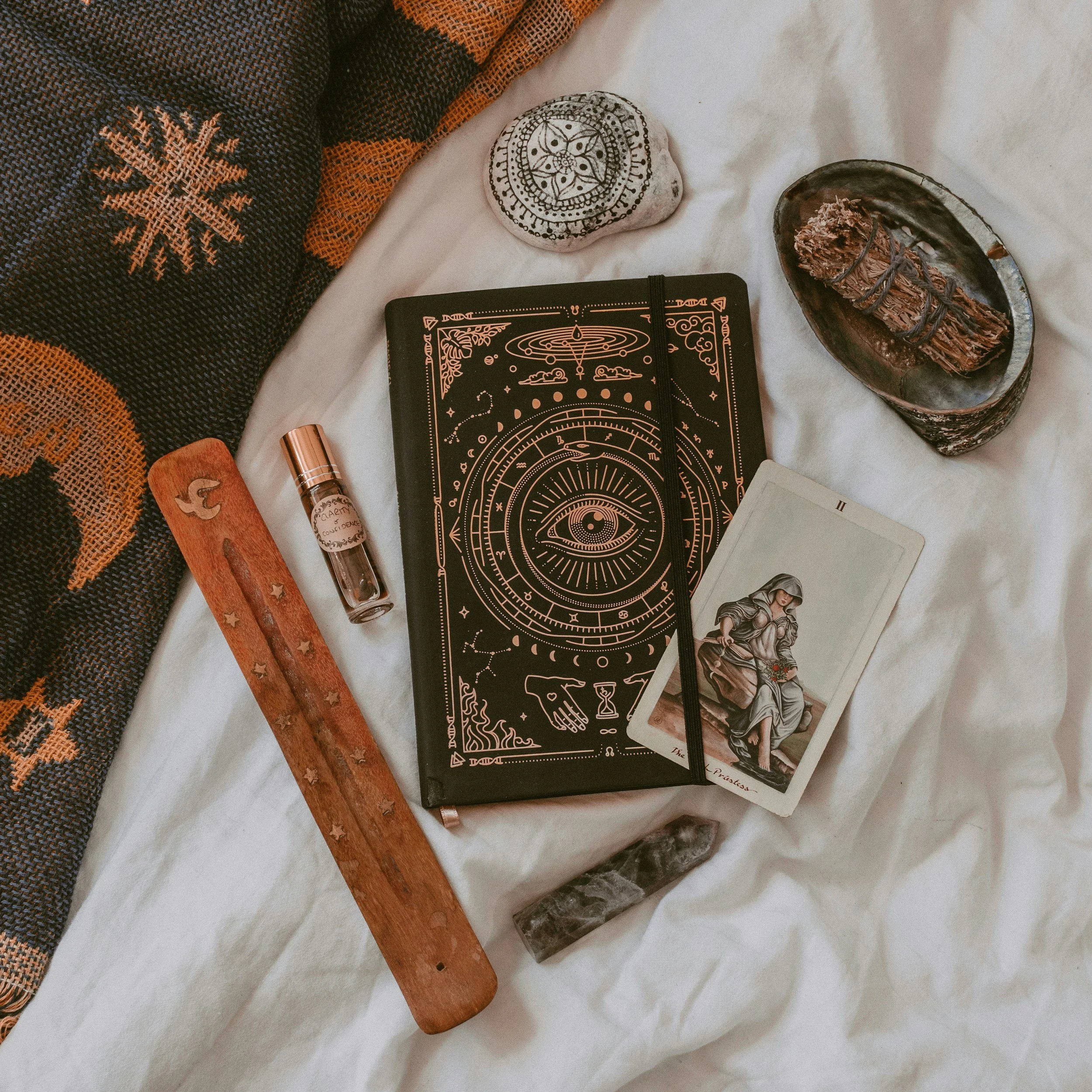 A collection of spiritual and mystical items on a white fabric surface, including a black notebook with gilded magical symbols, a tarot card featuring a seated woman, a mineral stone, a small vial, a wooden pendulum with star and moon cut outs, a black and silver bowl with dried herbs or incense, and a patterned cloth with star-shaped designs.