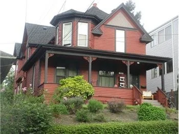 A two-story red wooden house with a front porch and steep roof, surrounded by greenery.