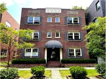 Brick three-story residential building with black awning over entrance, surrounded by green trees and shrubs, on a sunny day.