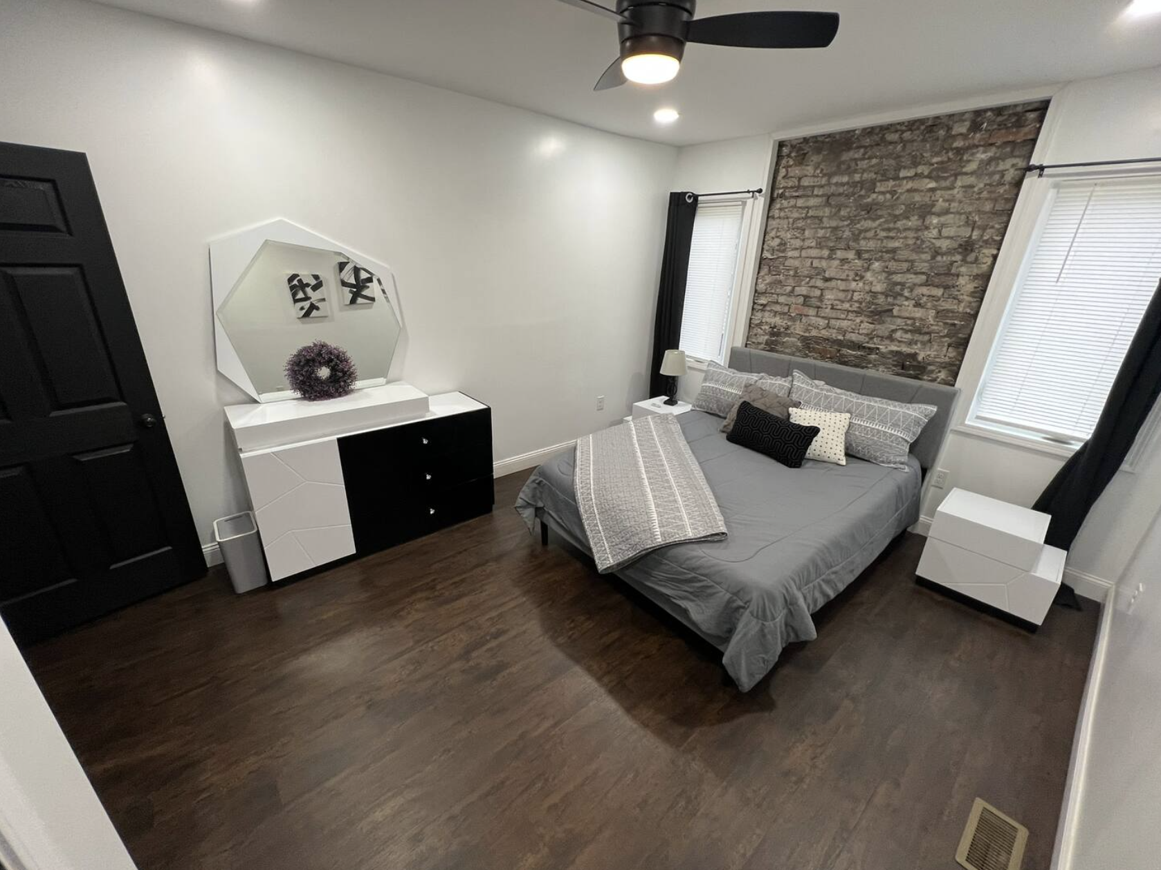 A bedroom with dark wood flooring, a bed with gray bedding and multiple pillows, two windows with blinds and black curtains, a nightstand with a lamp, a dresser with a large mirror and decorative purple flower, and an exposed brick wall.
